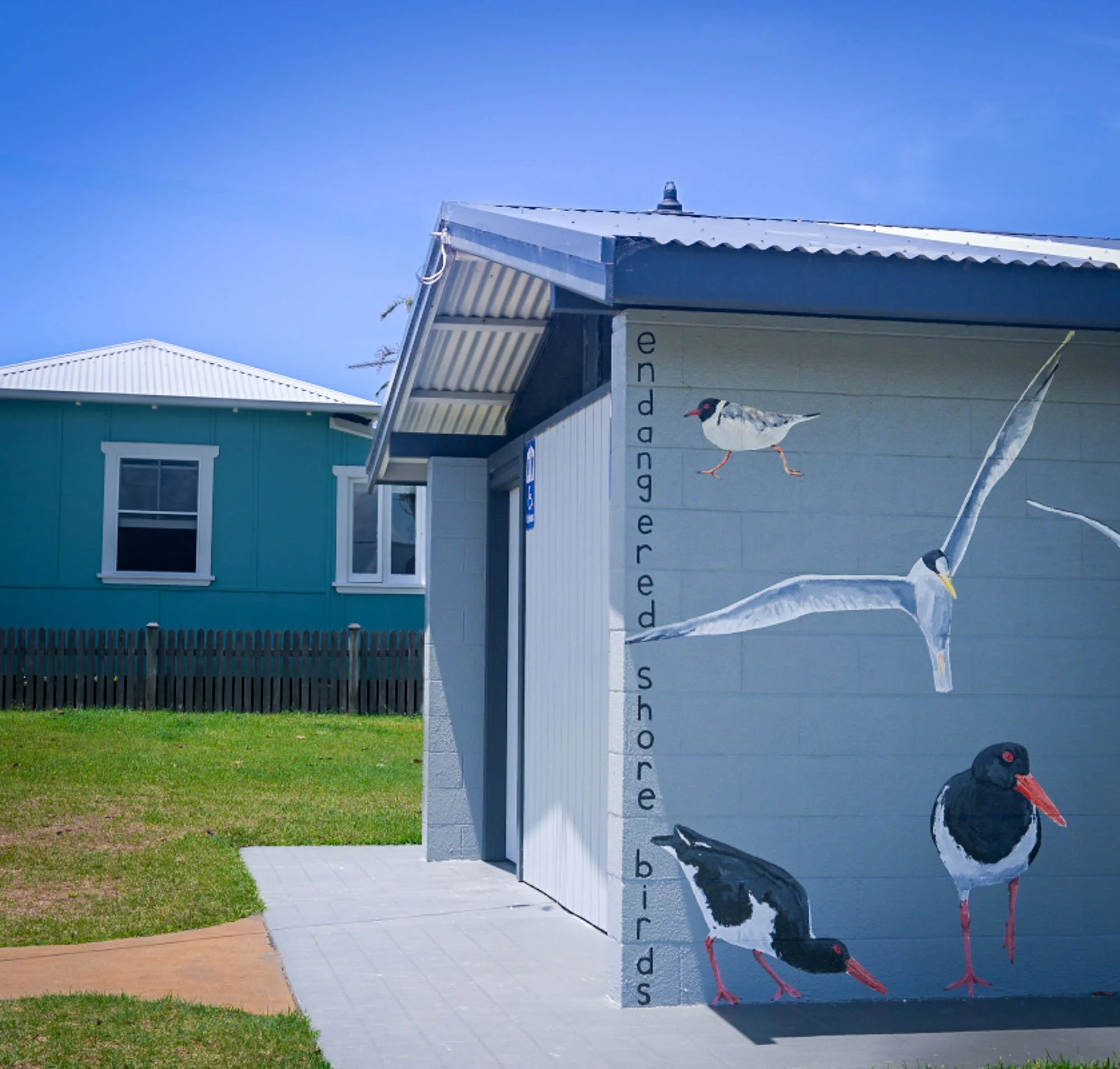 A house with light blue walls and white trim, featuring a window and door, is visible in the background. In front, there is a white, rectangular mailbox with black and red illustrations of a bird, and large, vertical, black text spelling out 'bird fe