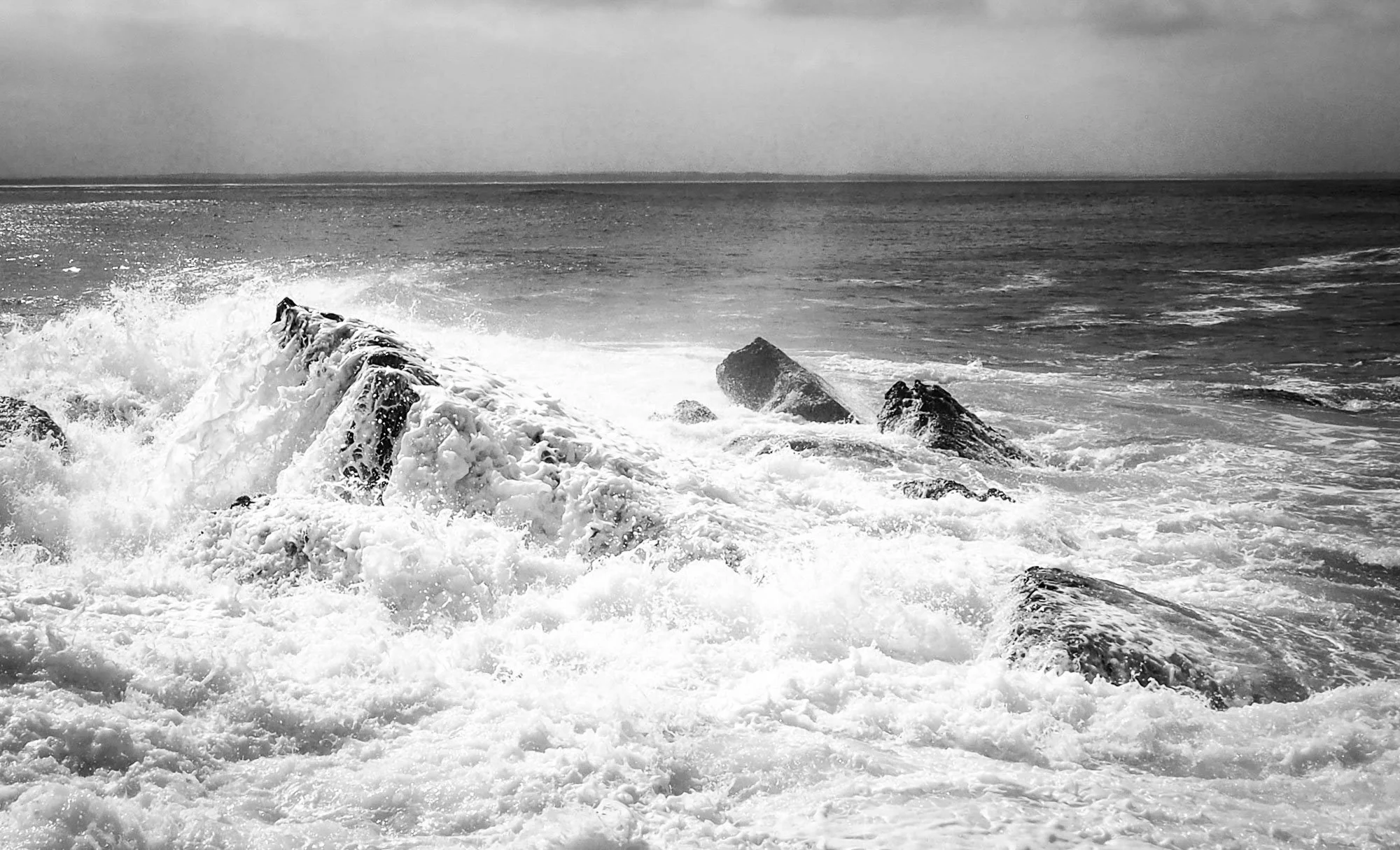 Black and white photo of ocean waves crashing against rocks near the shoreline.