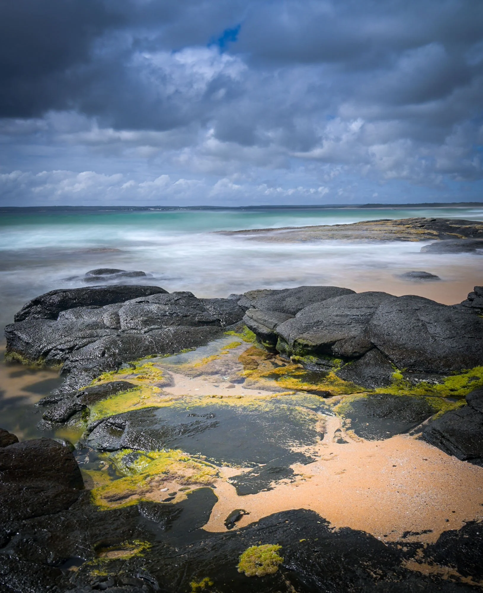 A rocky shoreline with dark rocks covered in green seaweed, sandy beach, and waves crashing against rocks under a cloudy sky.