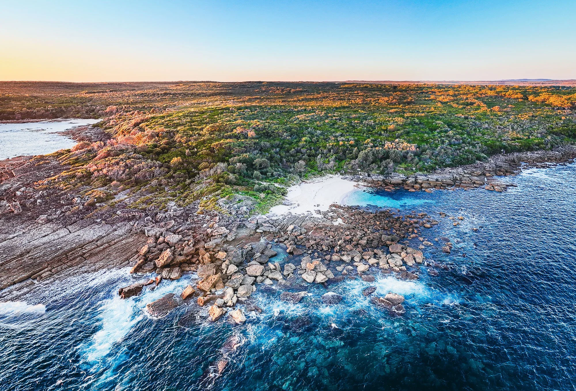 Aerial view of rocky coastline with dense green and pink flowering shrubbery, a small sandy beach, and clear blue ocean water, under a clear blue sky at sunset or sunrise.