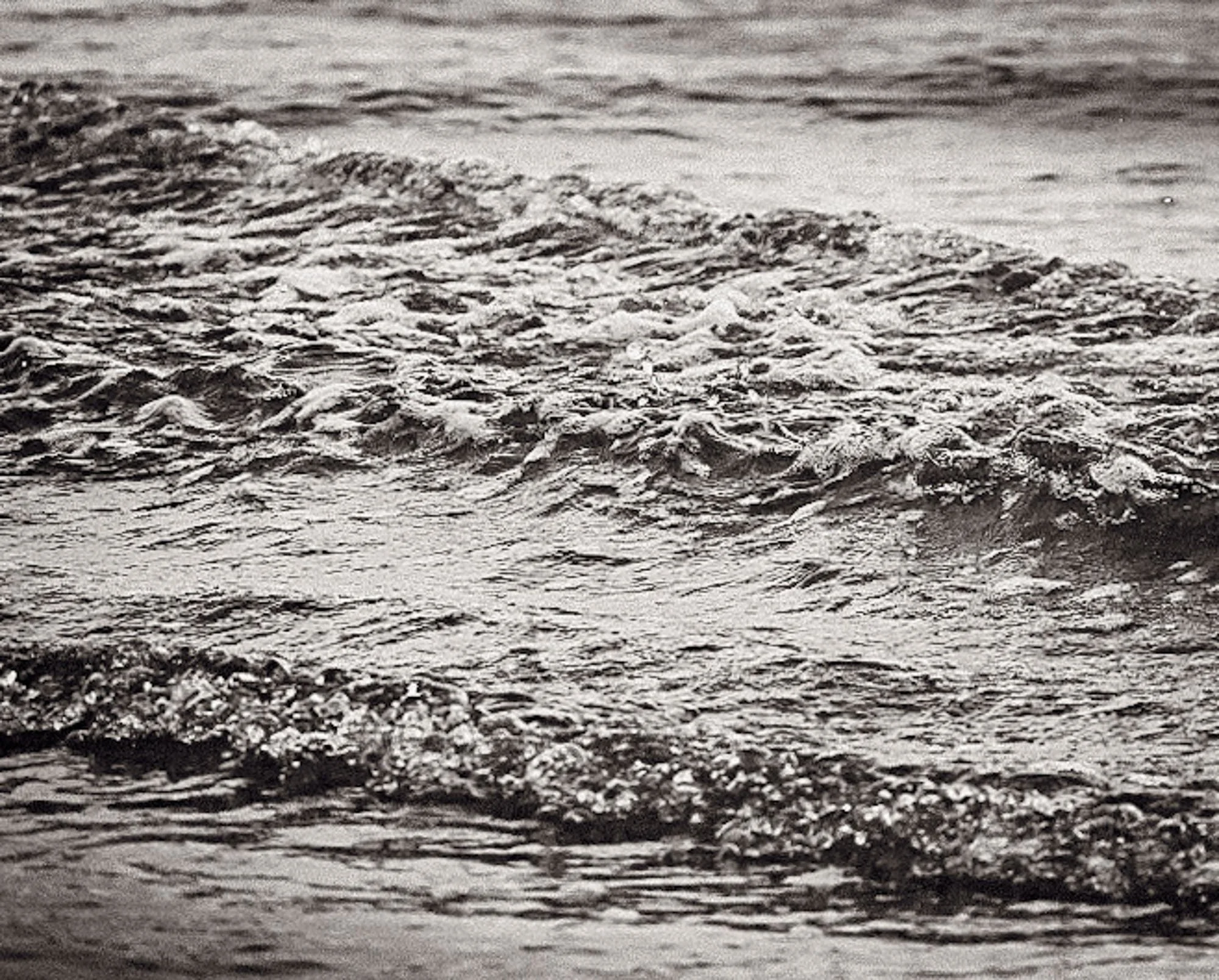 Black and white photograph of ocean waves gently crashing onto a pebbled beach.