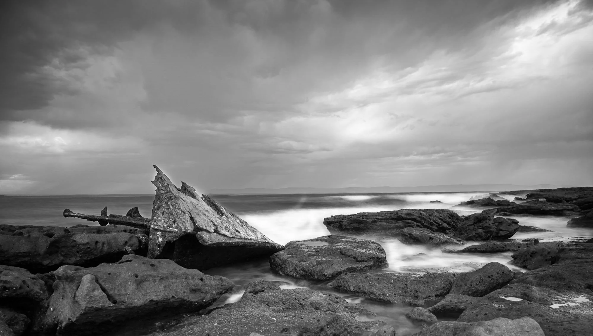 Black and white photograph of a rocky shoreline with a large, jagged piece of driftwood in the foreground, rough ocean waves, and a cloudy sky overhead.