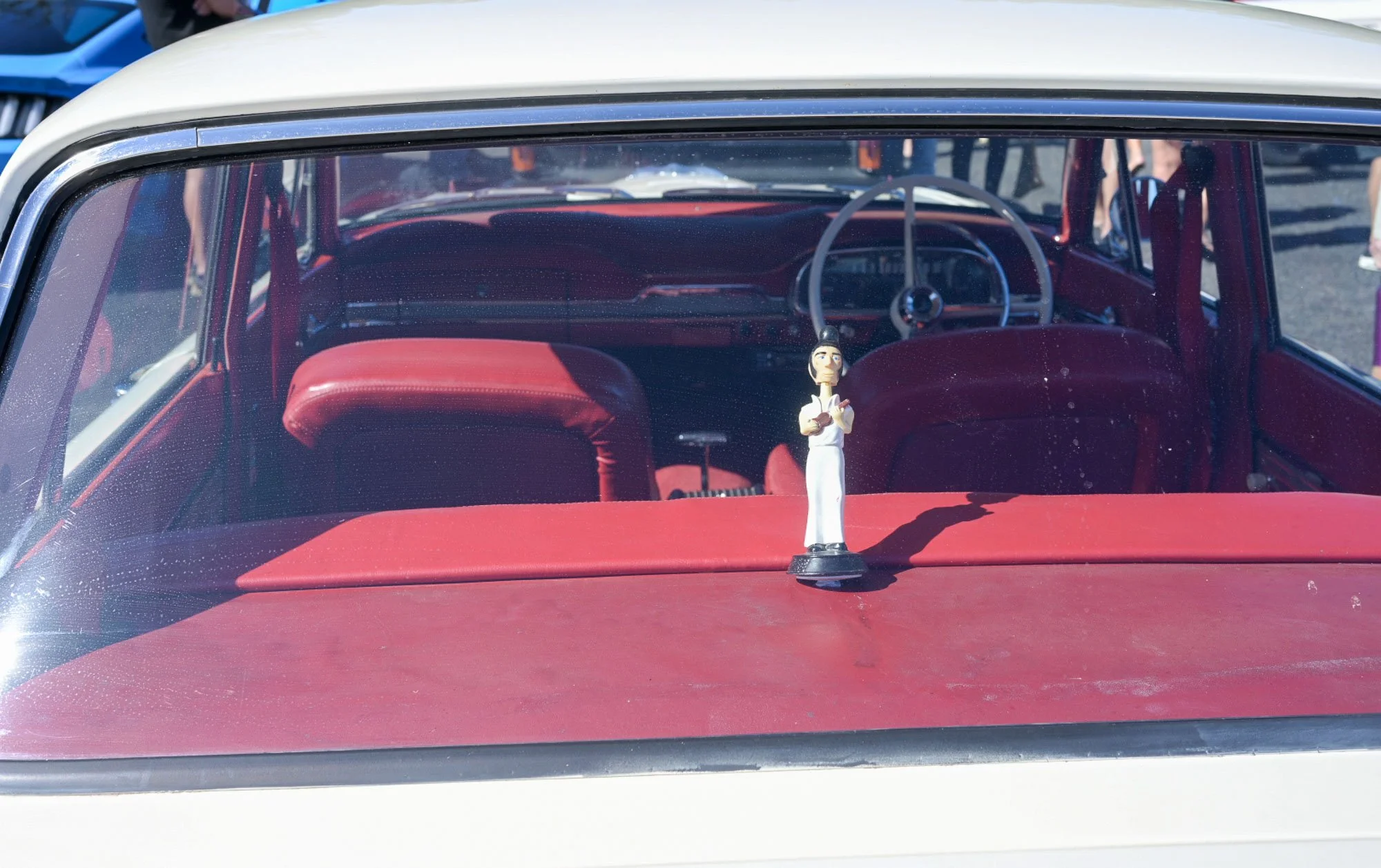 A vintage car with a red interior and a black steering wheel. There is a small figurine of a man dressed in white, playing a guitar, placed on the dashboard. The photo is taken from outside the car, looking through the windshield.