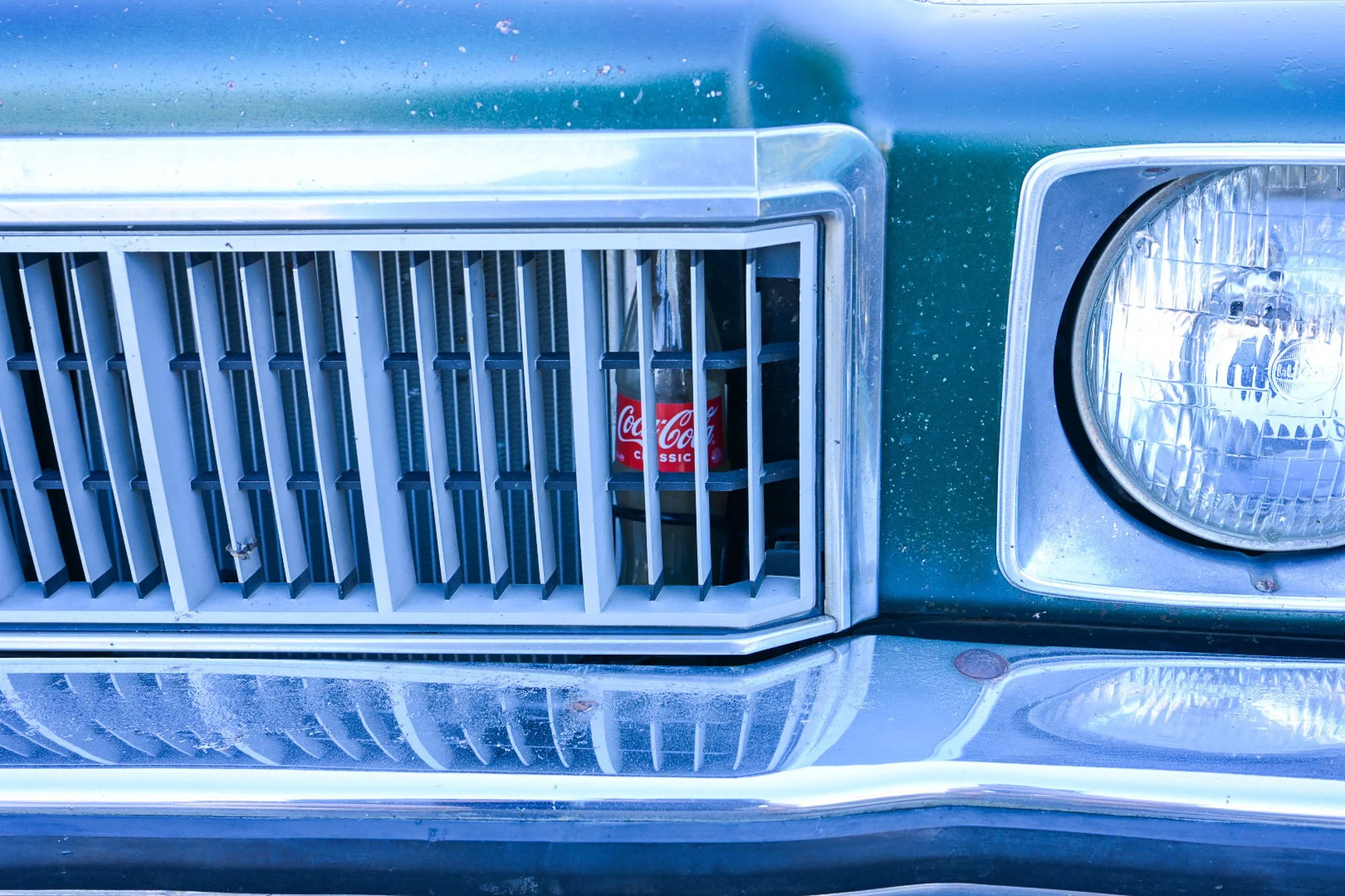 A vintage car with a blue-green body and chrome accents, featuring a front grille with vertical slats, a headlight with a chrome surround, and a can of Coca-Cola Classic secured behind the grille.