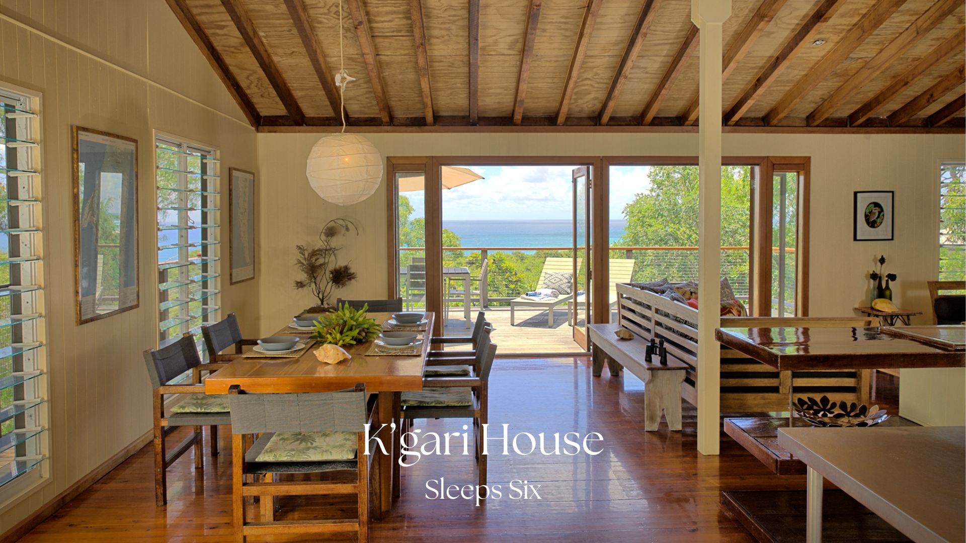 Interior view of a house with a dining area and living space, open to a balcony with ocean view, including a dining table, chairs, artwork, and outdoor furniture.