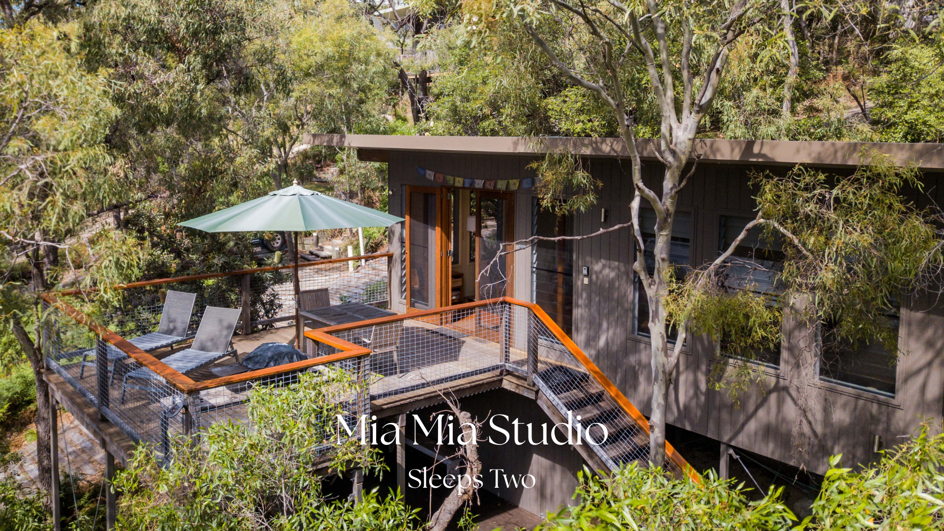 Aerial view of a small, modern house with a large outdoor wooden deck surrounded by trees, featuring a green patio umbrella, lounge chairs, and a cozy seating area.
