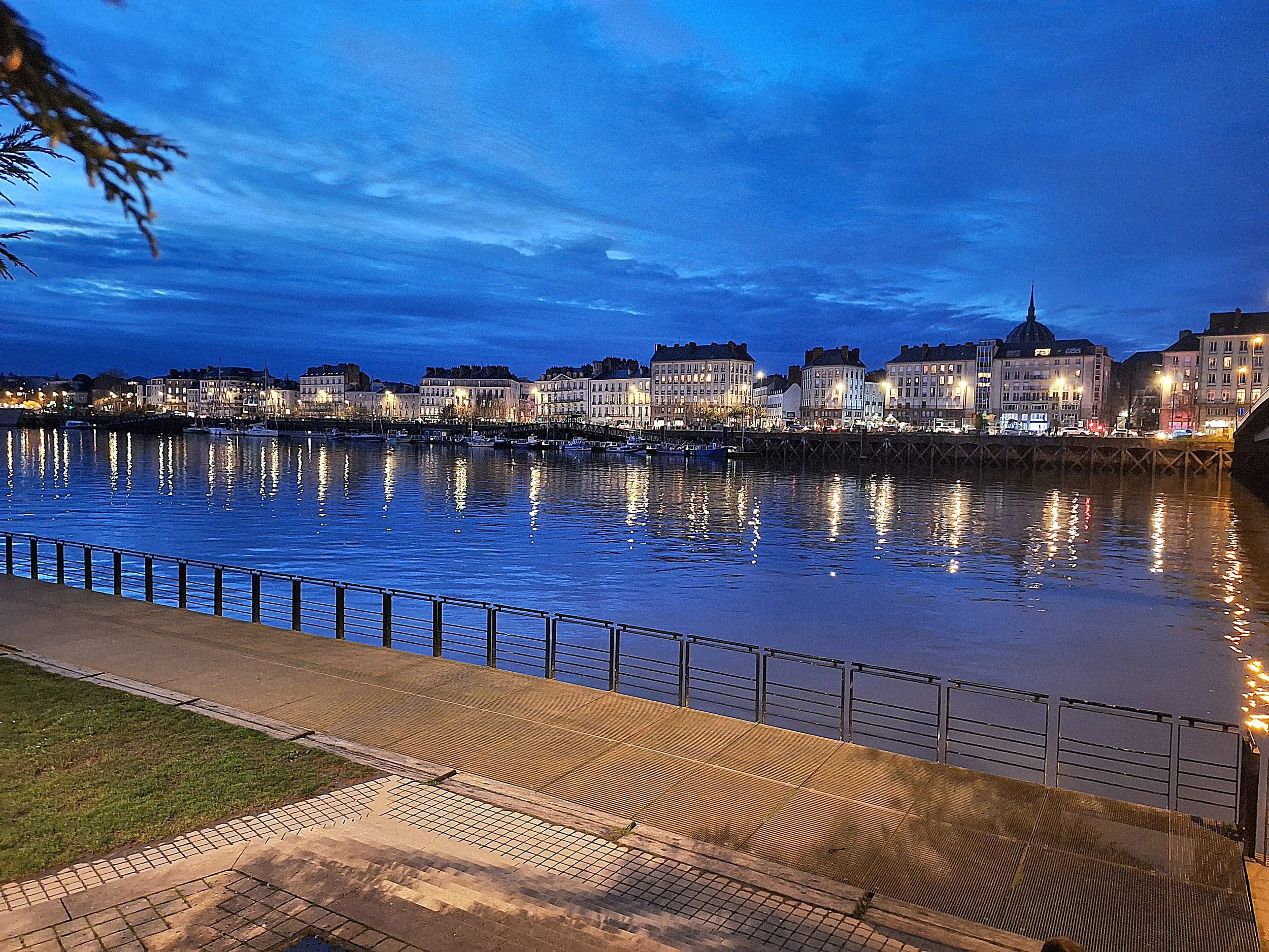 Un paysage urbain au bord d'une rivière la nuit avec des bâtiments éclairés et un ciel bleu foncé.