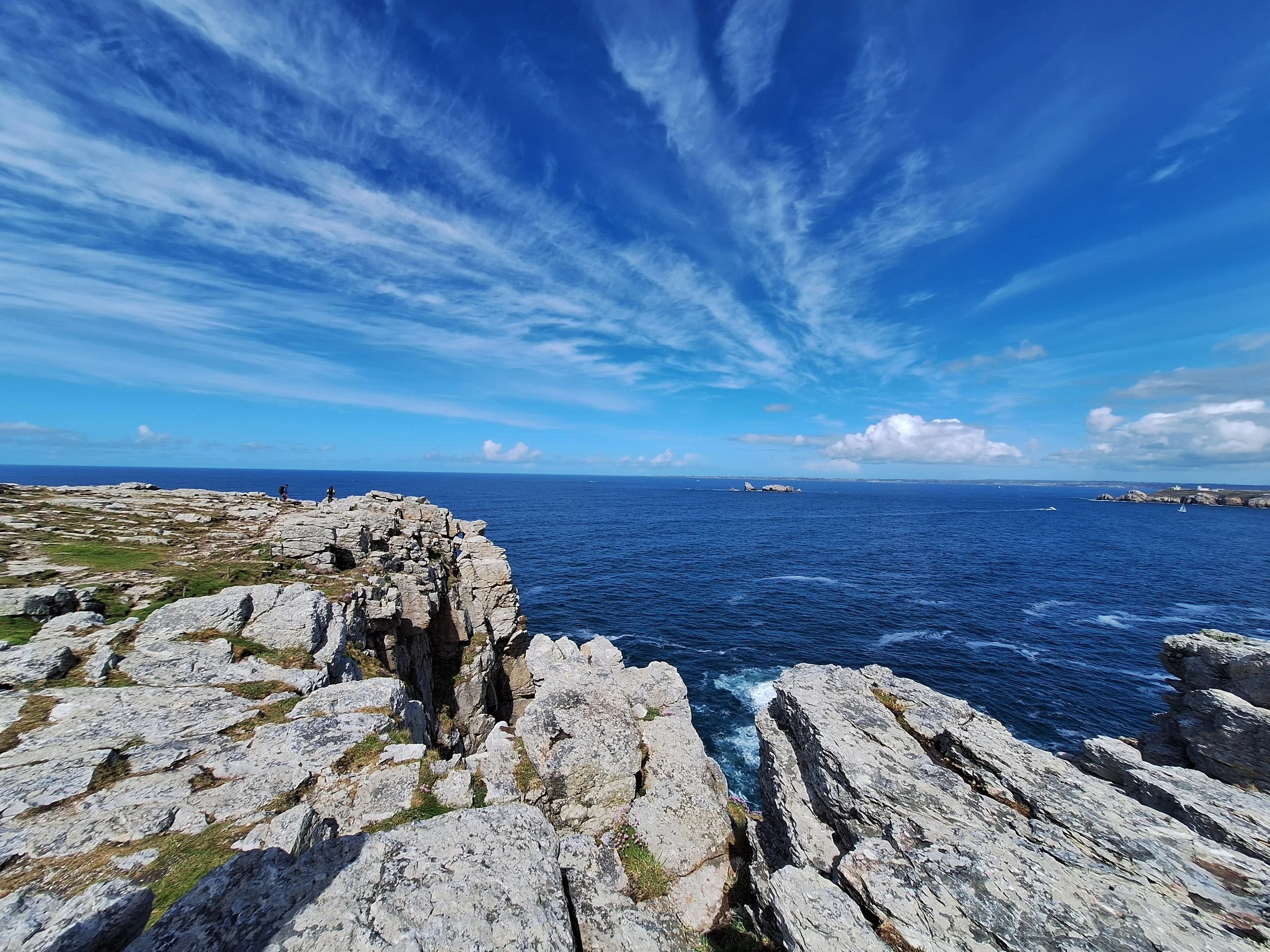 Côte rocheuse avec vue sur la mer sous un ciel bleu avec des nuages blancs.