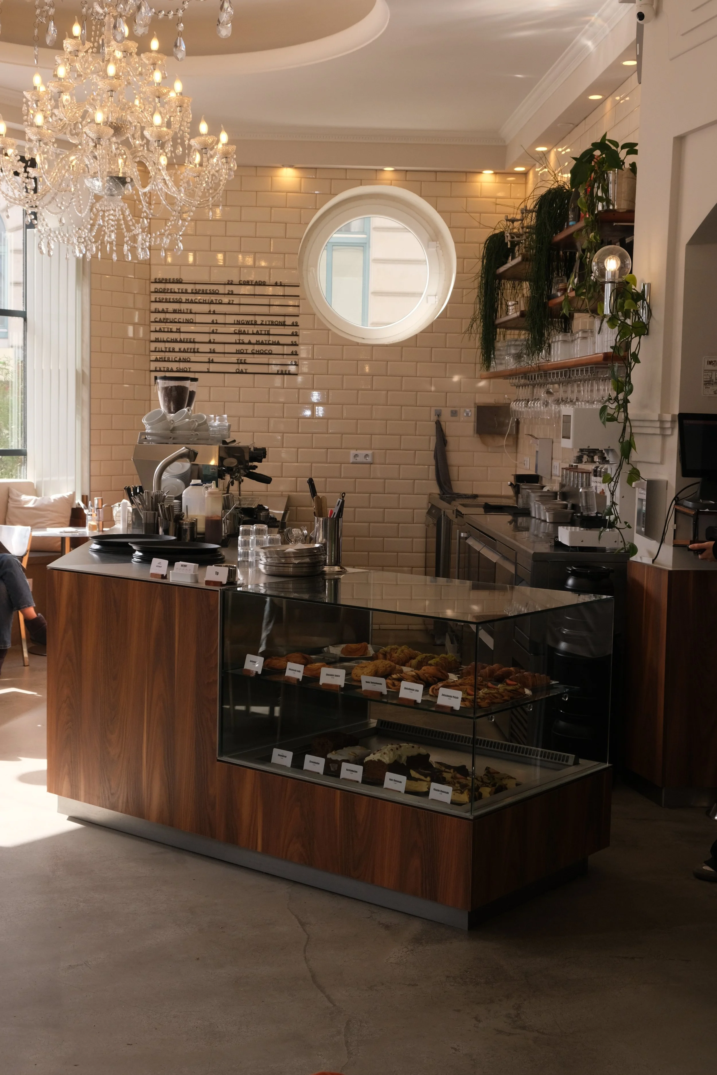 Innenaufnahme eines Cafés mit einer Theke, auf der Kekse und Kuchen in einer Glasvitrine präsentiert werden, einem Kaffeeautomat, Tassen, Gläser und Besteck. Im Hintergrund eine an der Wand montierte Speisekarte und Dekoration mit Pflanzen und einem 