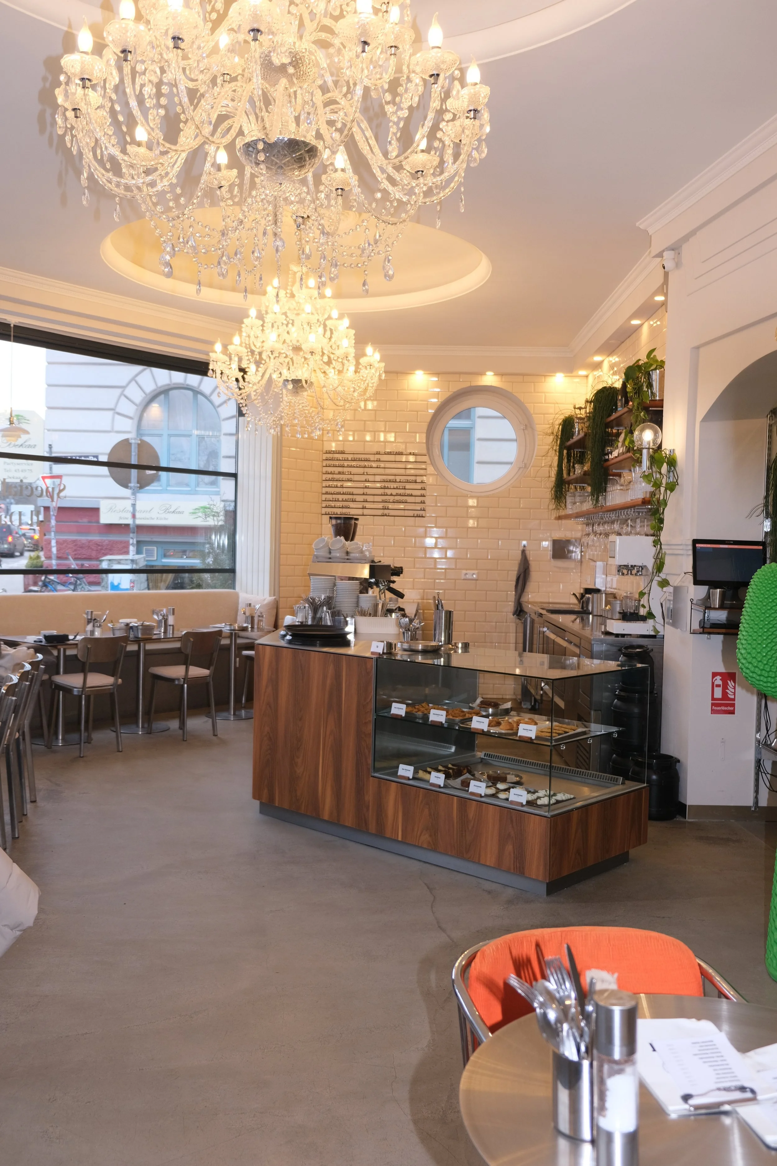 Innenansicht eines elegant eingerichteten Cafés mit Kronleuchtern, einer Theke mit Kuchen und Kaffeemaschine, großen Fenstern und gemütlichen Sitzgelegenheiten.
