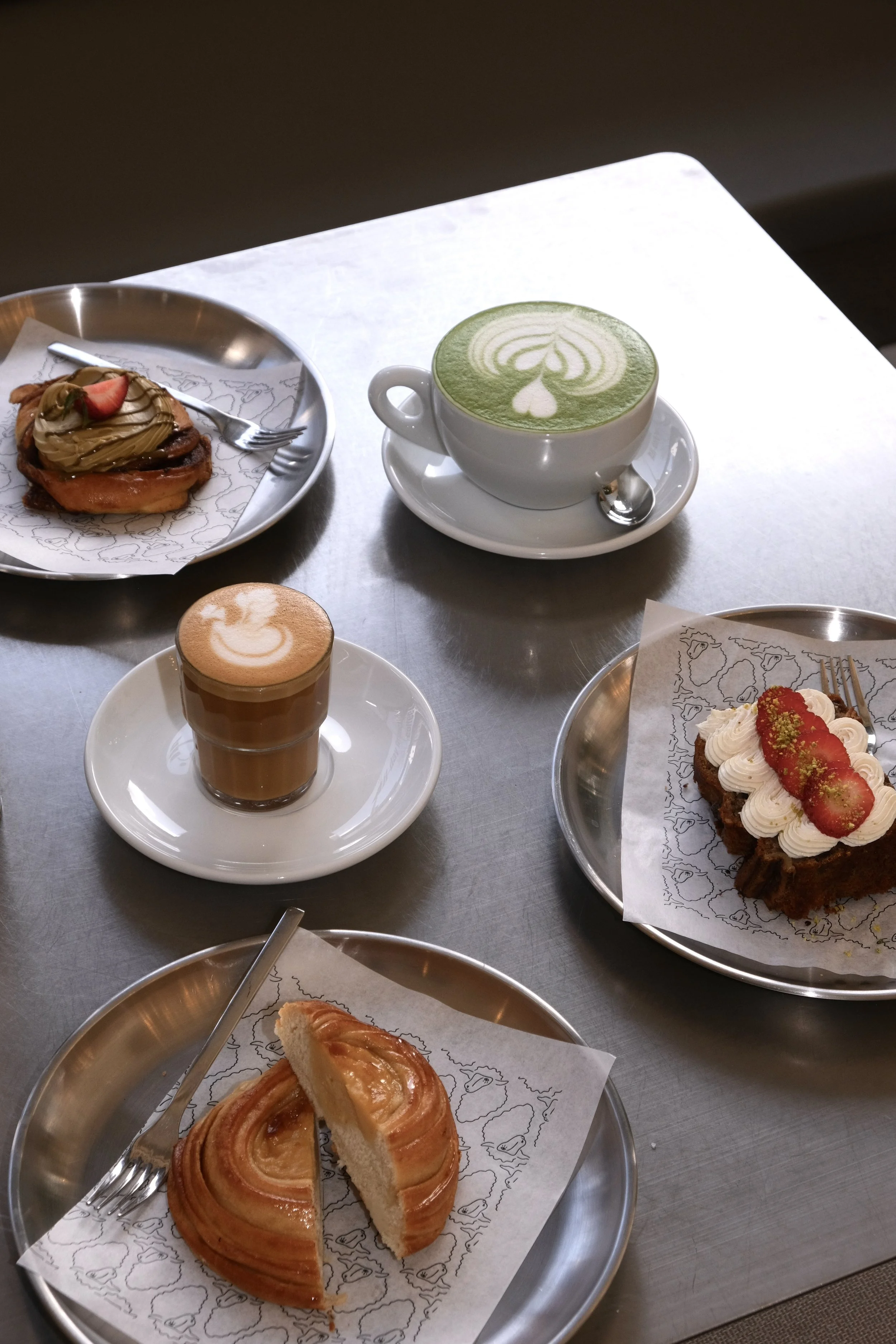 Ein Tisch mit verschiedenen Desserts, darunter ein Matcha-Latte mit Latte-Art, ein Stück Apfelkuchen, ein Cappuccino mit Milchschaum und ein Stück Käsekuchen mit Erdbeeren.