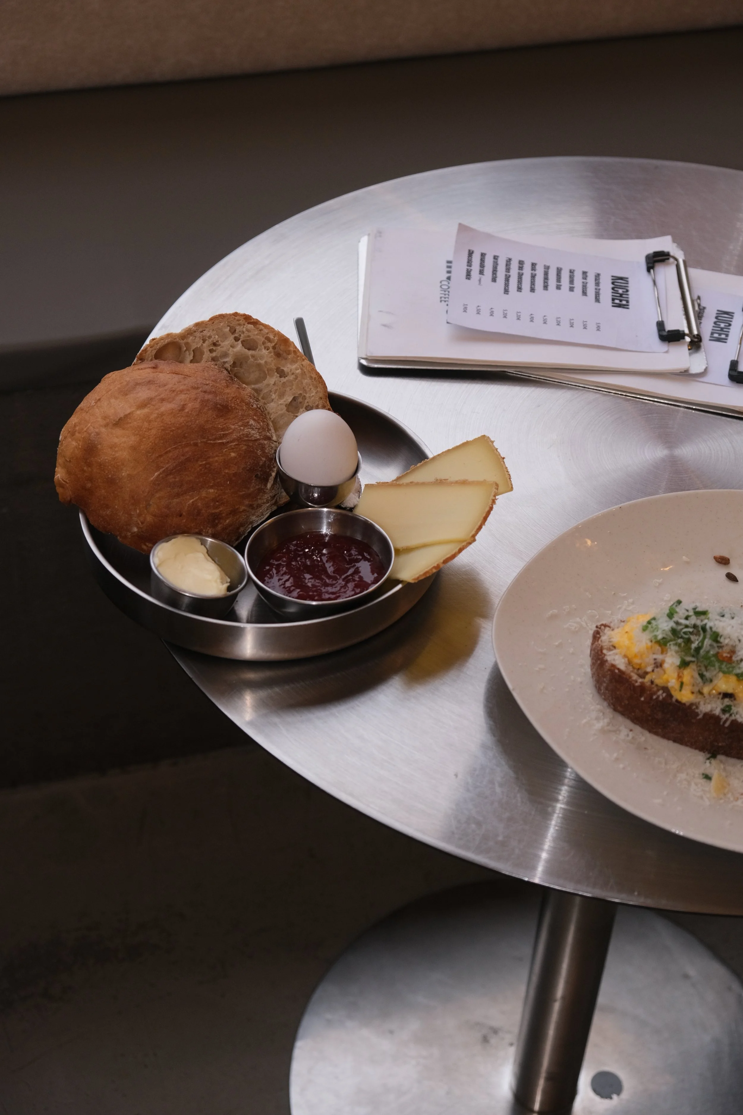 Frühstücksteller mit Brötchen, hartgekochtem Ei, Käse, Marmelade, Butter und einer Scheibe Brot mit Ei und Kräutern, auf einem runden Metalltisch.