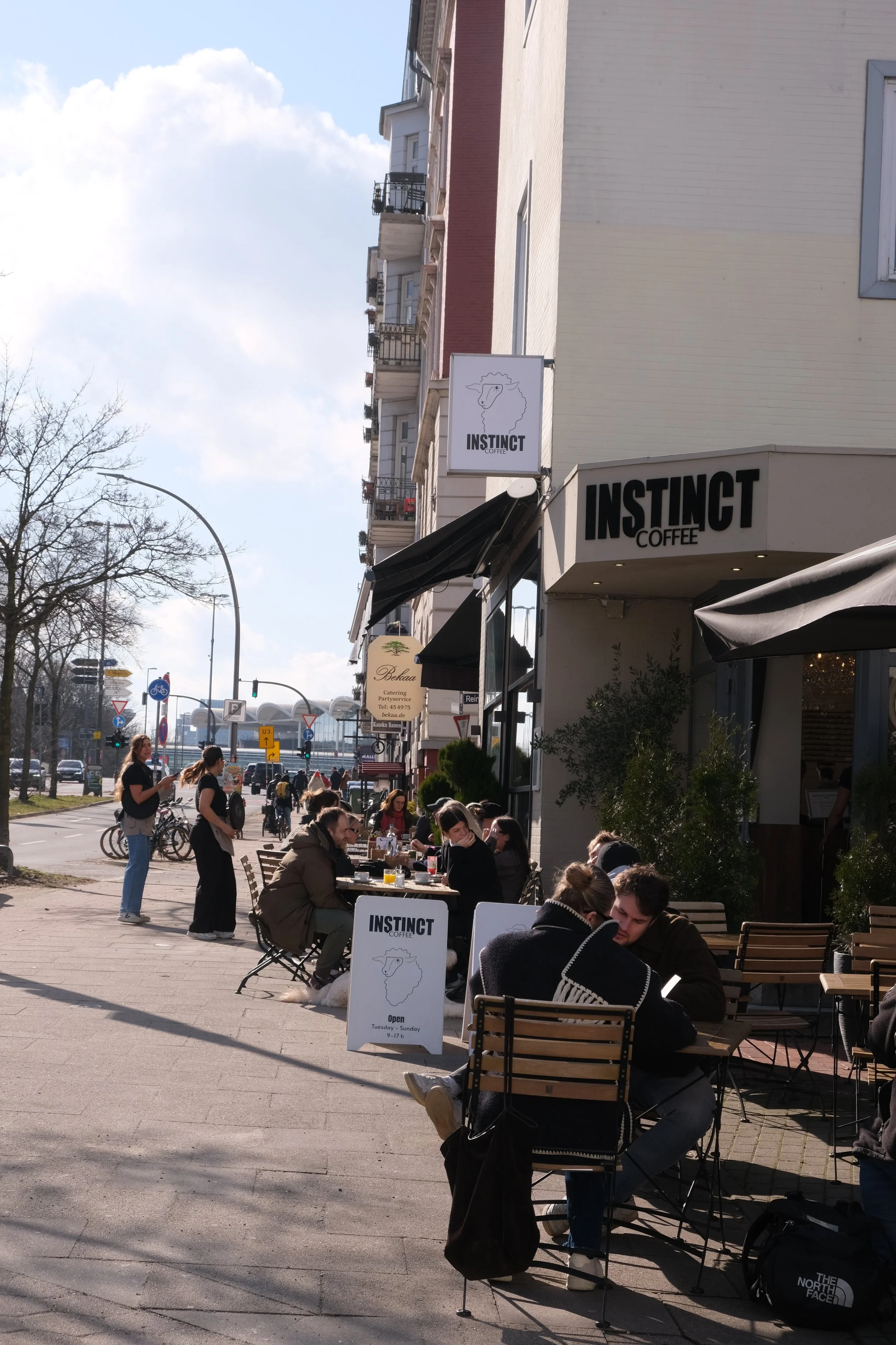 Straßencafé im Sonnenschein mit Menschen, die Getränke genießen. Ein Schild mit dem Logo 'INSTINCT COFFEE' ist sichtbar.