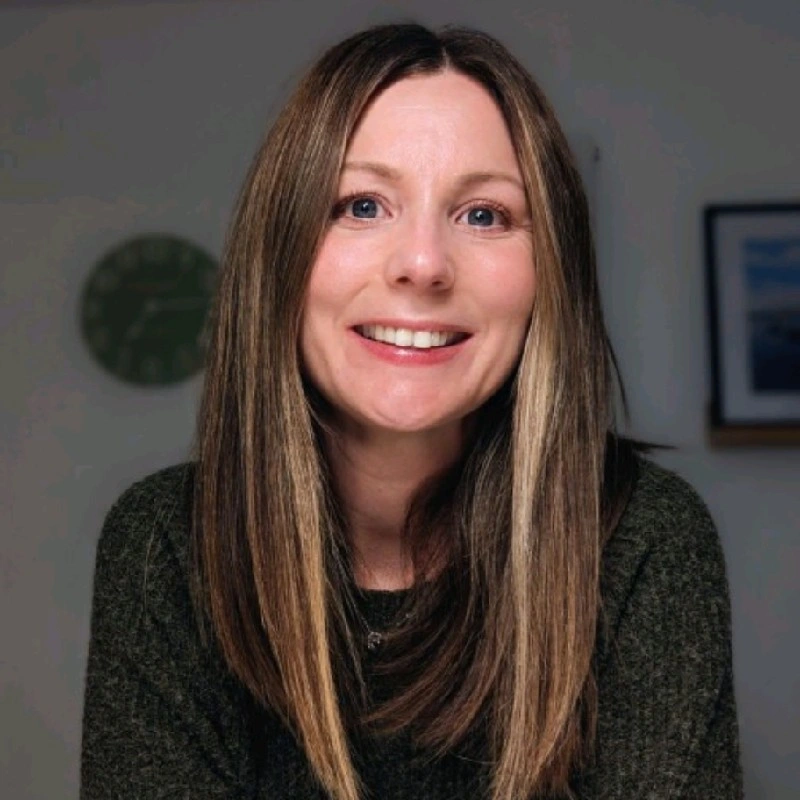 A smiling woman with blonde wavy hair sitting at a desk, holding a pen and writing in a notebook, with a coffee mug and a calculator on the desk, and a plant and books on a shelf in the background.