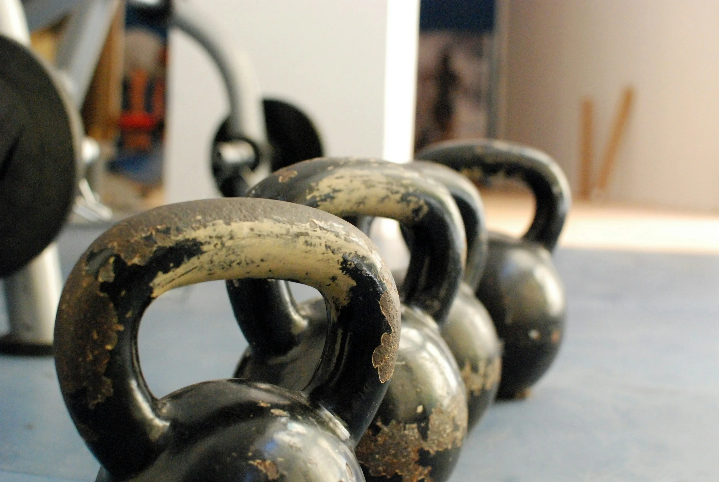 Three worn kettlebells on a gym floor with a dumbbell visible on the side in the background.
