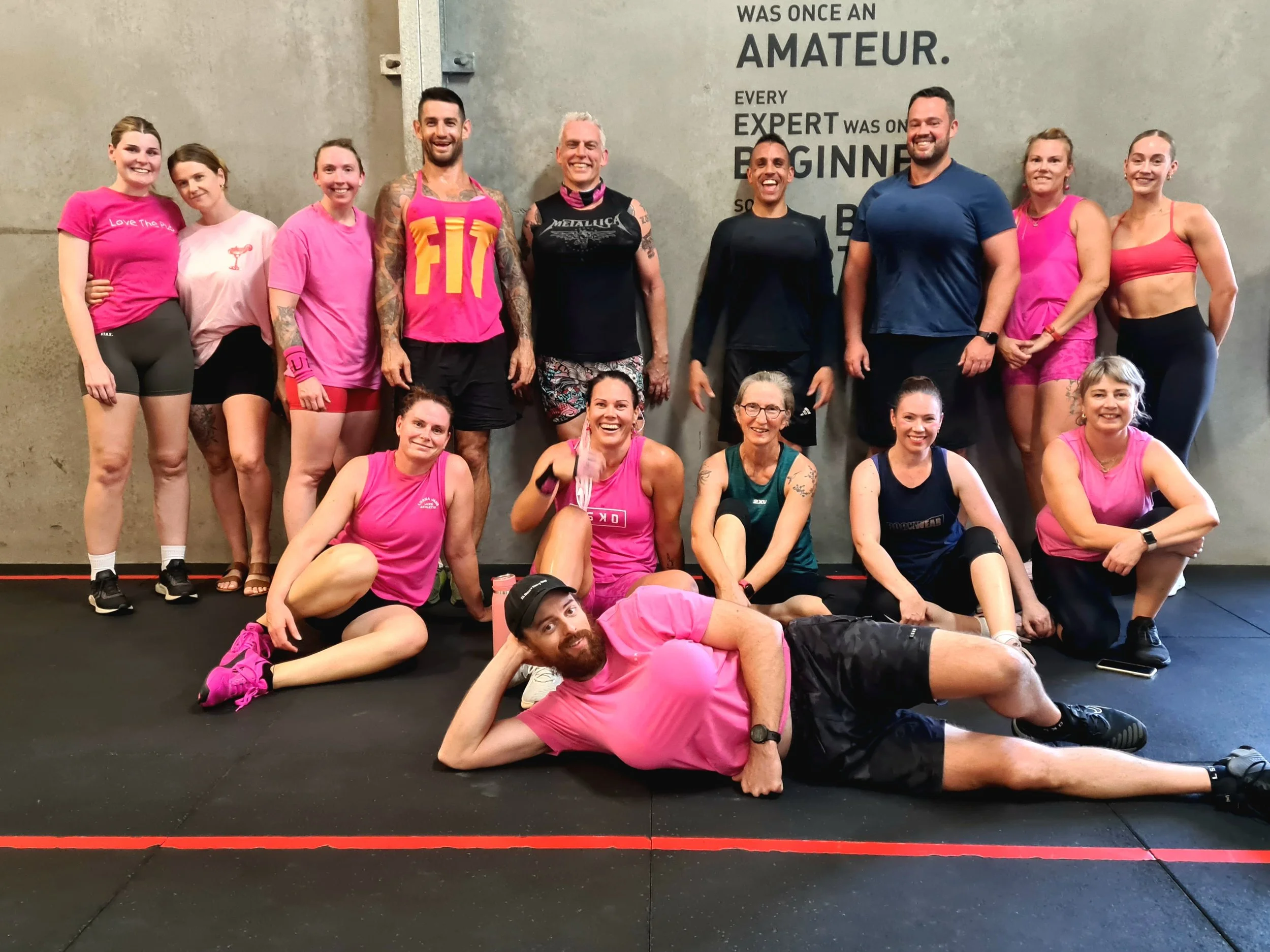 Group of people at the gym, some sitting, some standing, dressed in athletic wear, smiling for the camera. A man lies on the floor in front, wearing a pink shirt and black shorts. The wall behind has motivational text about amateurs and experts.