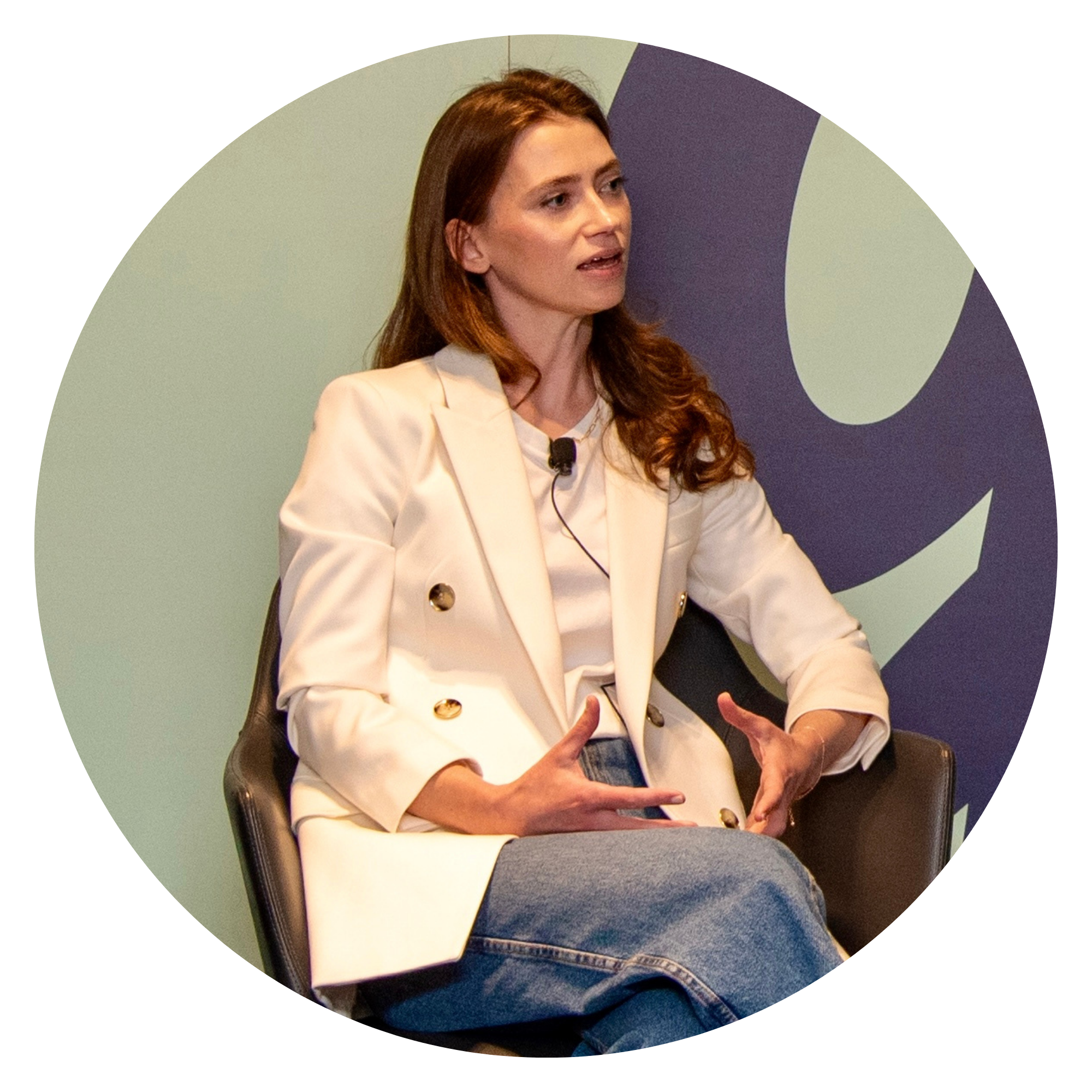 A woman is sitting at a table, speaking and gesturing with her hands. She is wearing a white blazer and a microphone attached to her shirt.