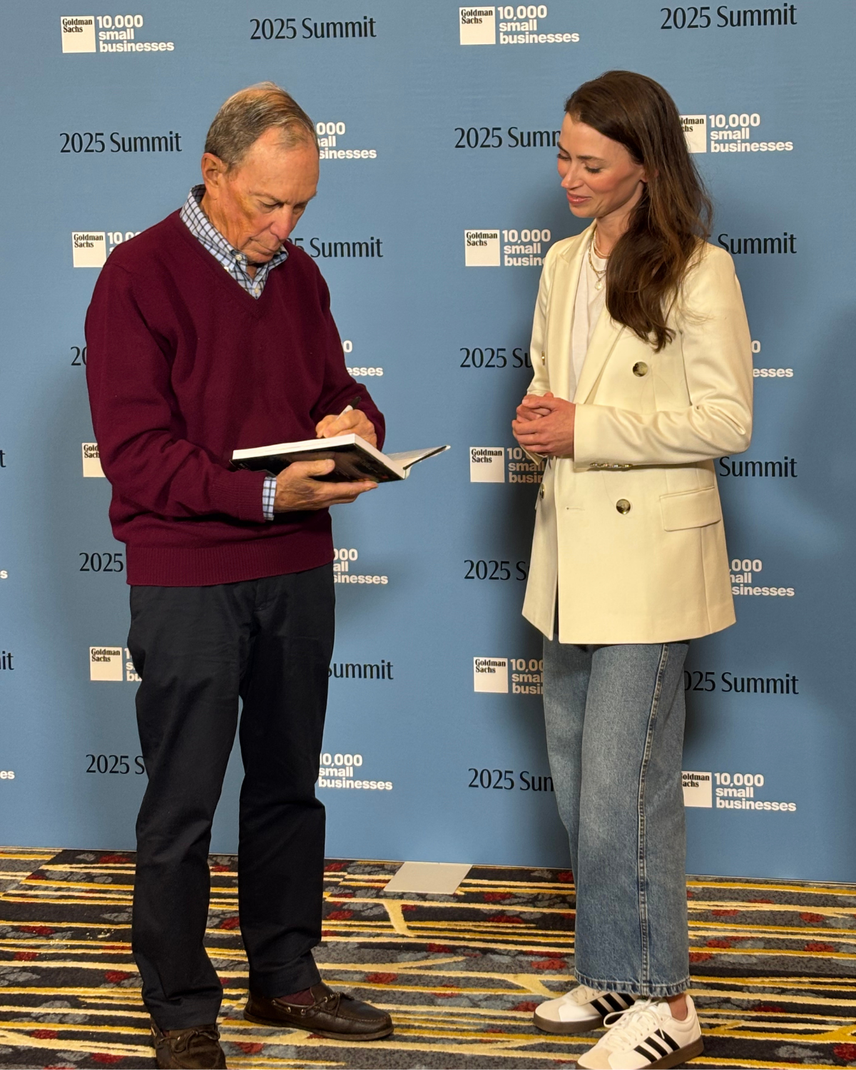 A man and woman are standing in front of a blue backdrop with '2025 Summit' and 'Goldman Sachs 10,000 small businesses' written on it. The man is looking down at a notebook, while the woman watches him, smiling softly. The man wears a maroon sweater over a checkered shirt and black pants. The woman wears a cream blazer, white shirt, blue jeans, and white Adidas sneakers.
