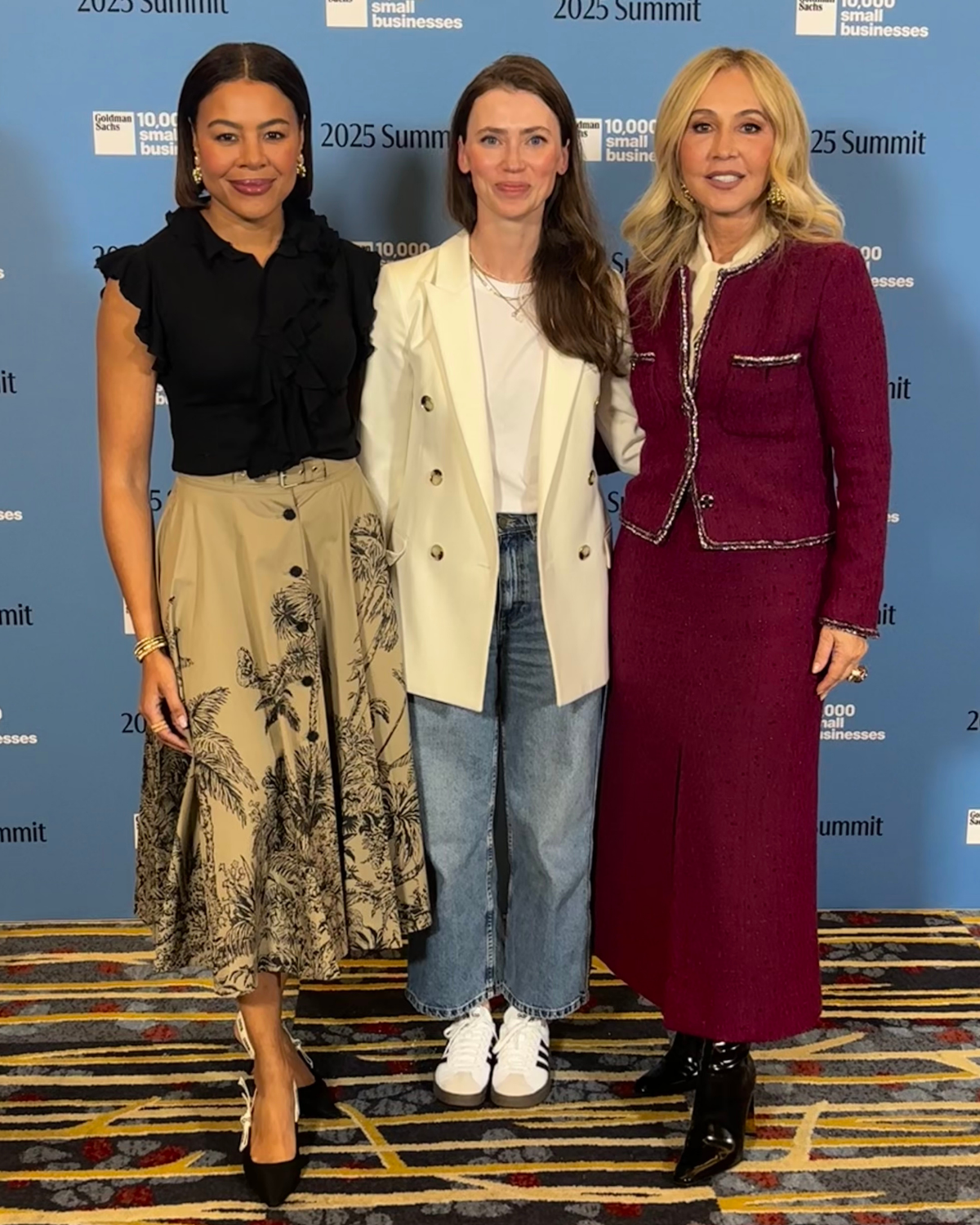 Three women standing in front of a blue backdrop with event logos, dressed in business casual attire, smiling for the camera.