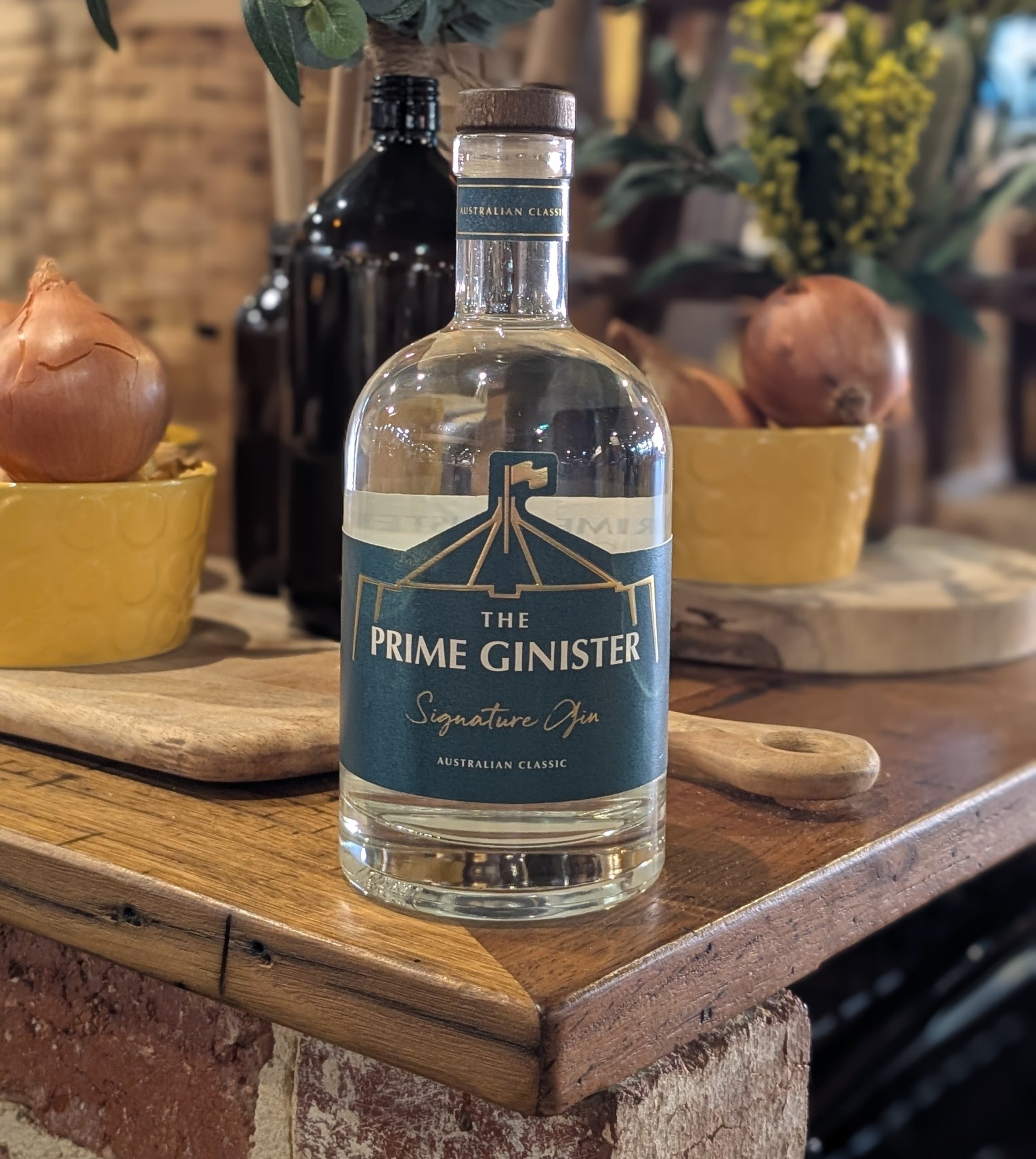 A clear bottle of The Prime Ginster Signature Gin with a label featuring a graphic of a flag on a pole. The bottle is placed on a wooden surface, with a background of onions in yellow bowls and other bottles and flowers.