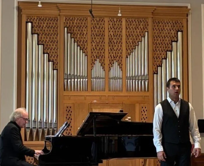 A pianist playing a grand piano and a singer standing in front of a large pipe organ in a concert hall