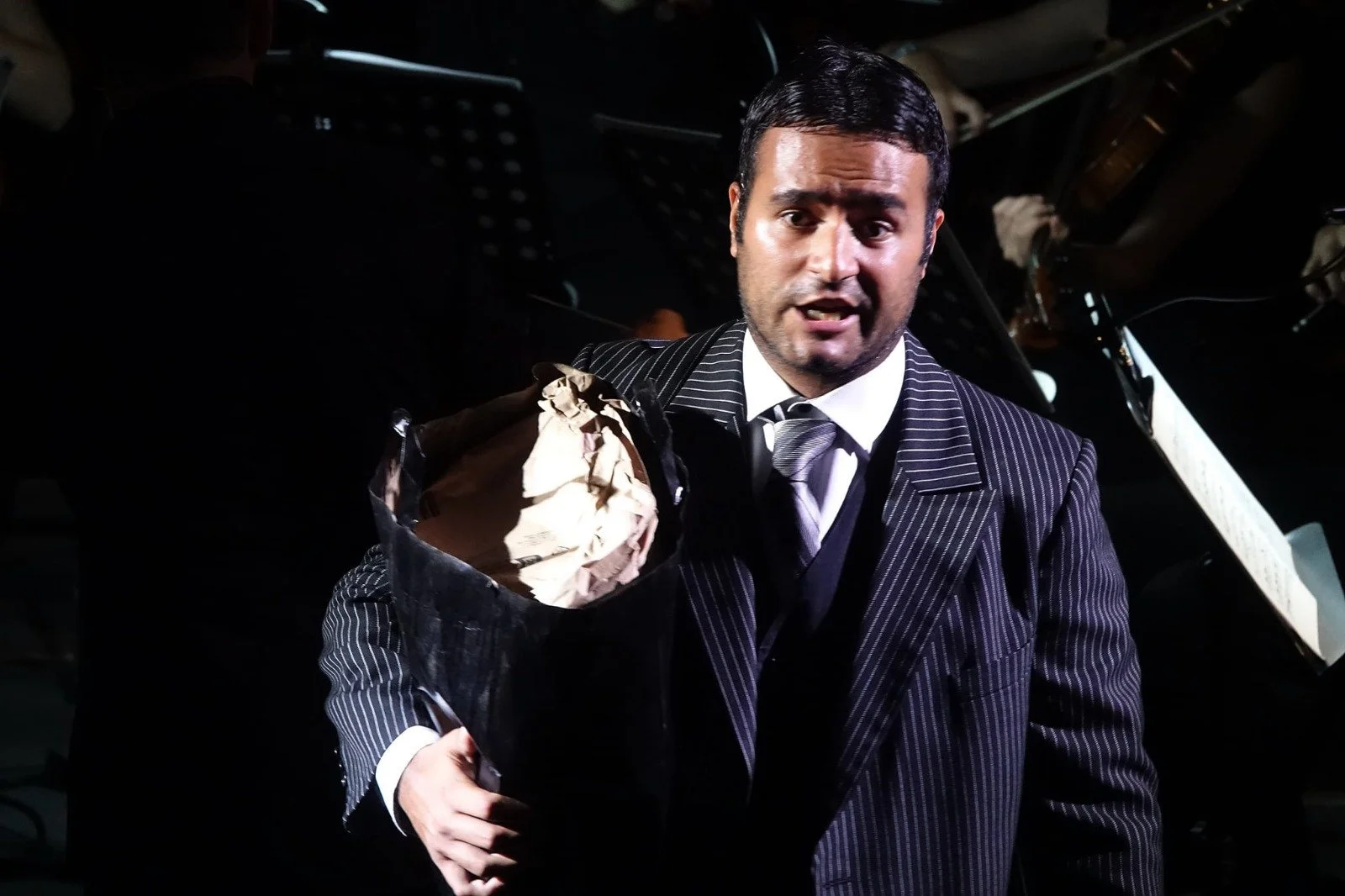 A man in a dark pinstripe suit and tie holding a trash bag with crumpled paper, standing in front of a grand piano.