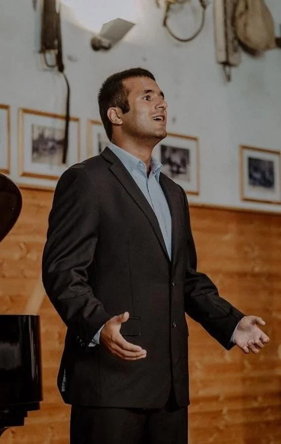 A man in a black suit and blue shirt is speaking or singing with a background of framed photos on the wall.