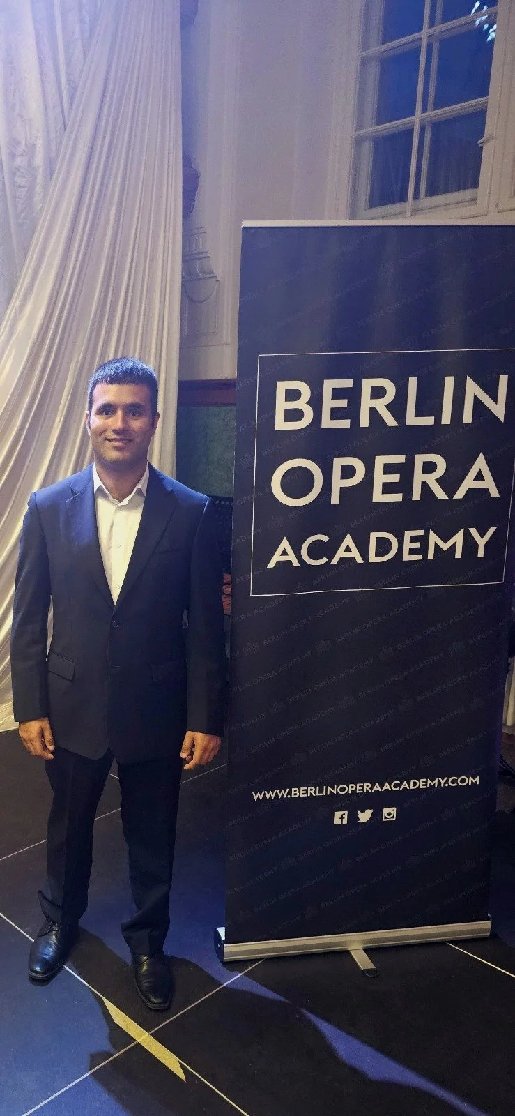 A man in a dark suit standing next to a banner that reads 'Berlin Opera Academy' at an event.