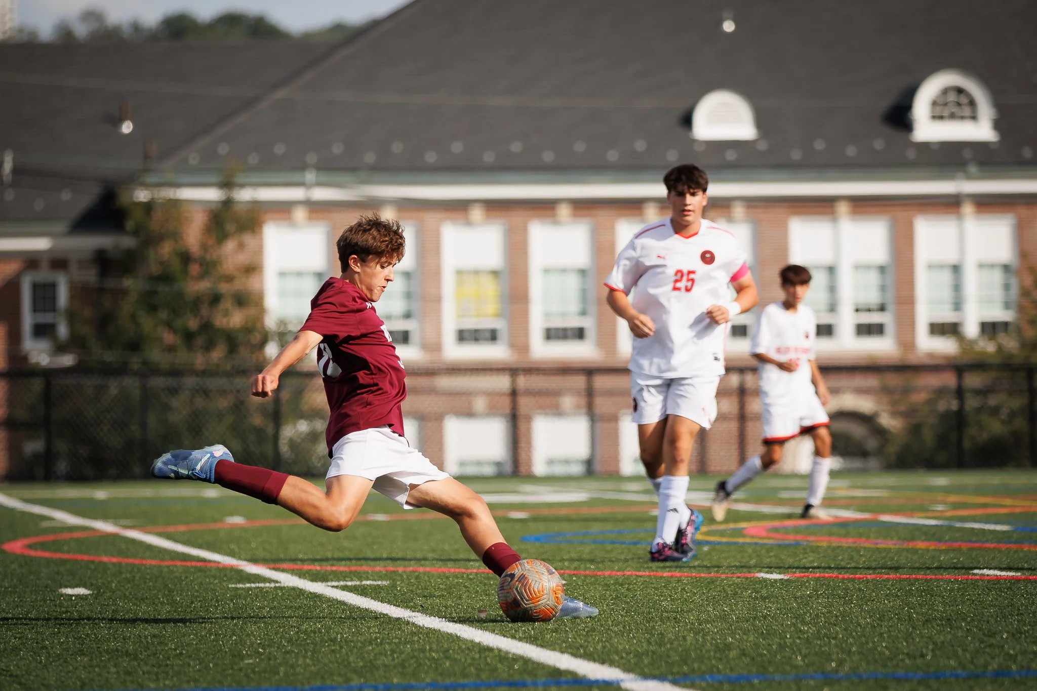 Young soccer player kicking a ball during a match on a green turf field with background of school building.