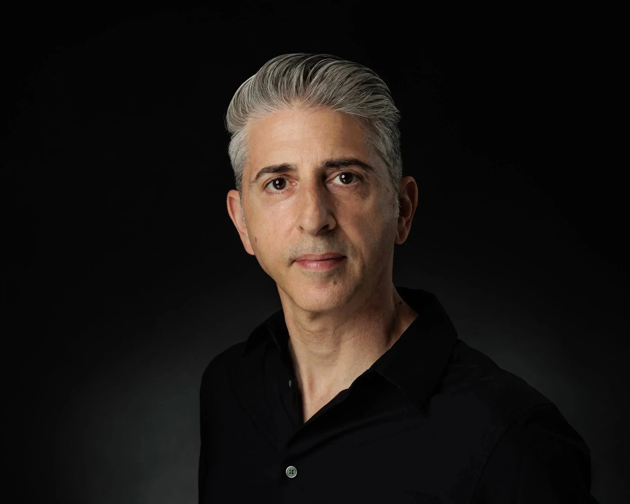Close-up portrait of a middle-aged man with silver hair, wearing a black shirt, against a black background.