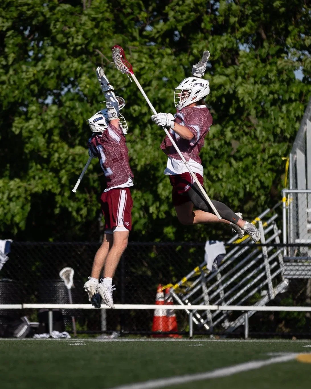 Two lacrosse players in maroon uniforms and helmets jump for the ball with their sticks crossed during a game on a field with green trees in the background.