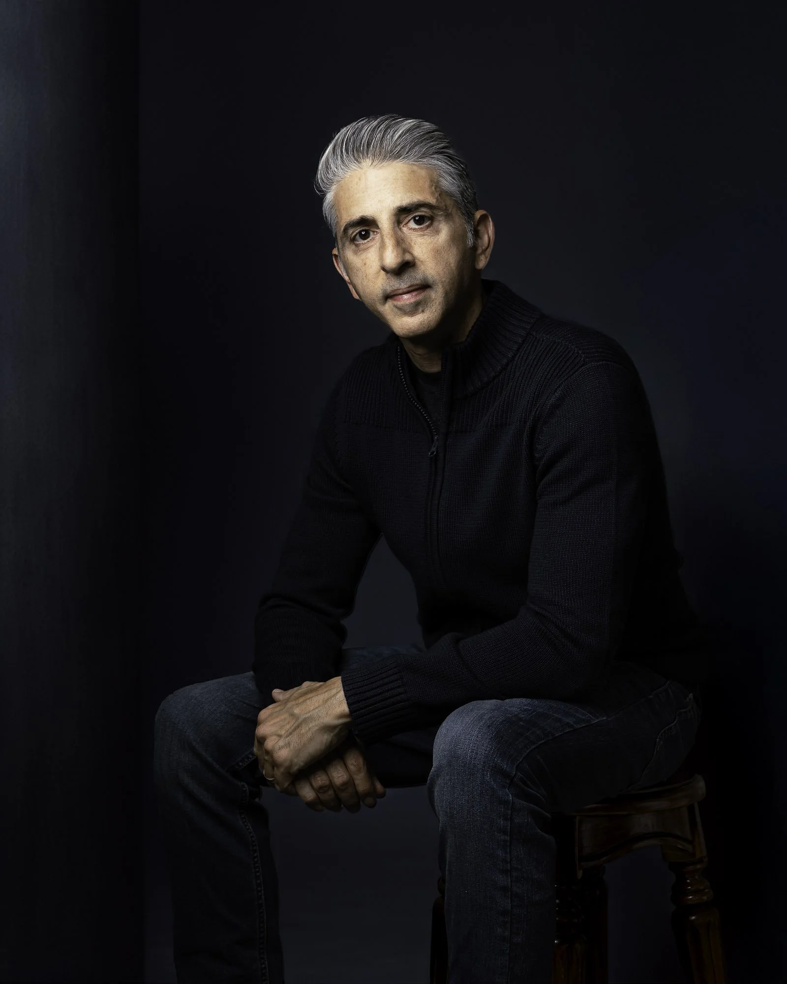 A middle-aged man with gray hair, wearing a black zip-up sweater and blue jeans, sitting on a wooden stool against a dark background.