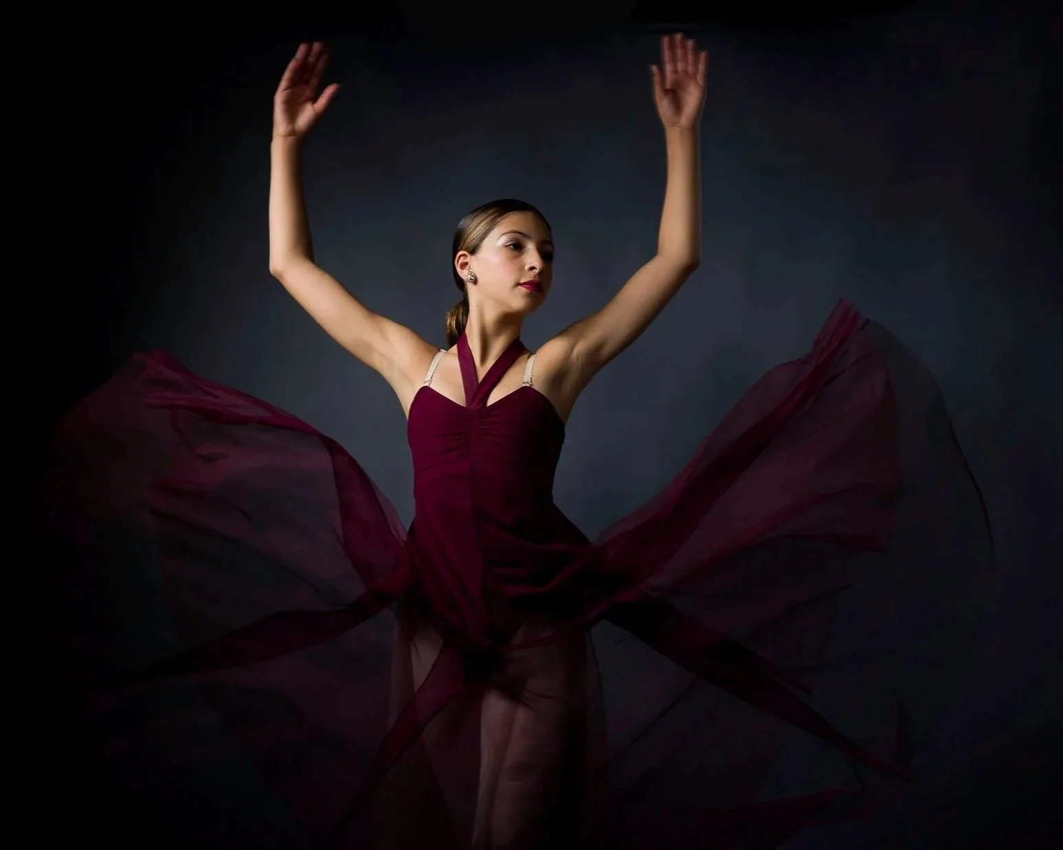 A ballerina in a maroon dress performing dance movements, with fabric flowing around her against a dark background.