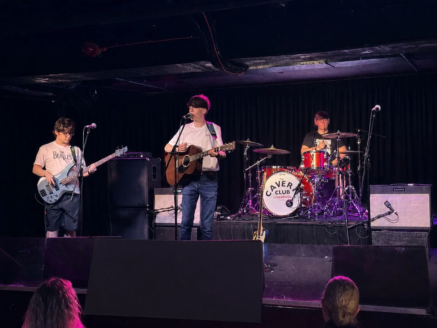 Three young boys performing on stage. One is playing bass guitar, one is singing and playing acoustic guitar, and the third is playing drums. The stage has black curtains and purple lighting, with an audience visible in the foreground.