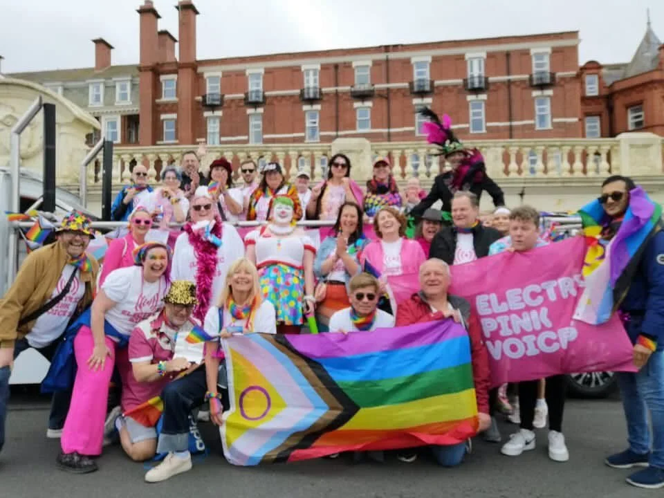 A group of people celebrating at a pride parade, holding rainbow flags and a pink banner that reads 'Electronic Pink Voice.' Many are wearing colorful clothing and accessories, with some dressed as clowns and others wearing rainbow or pink outfits, celebrating LGBTQ+ pride.