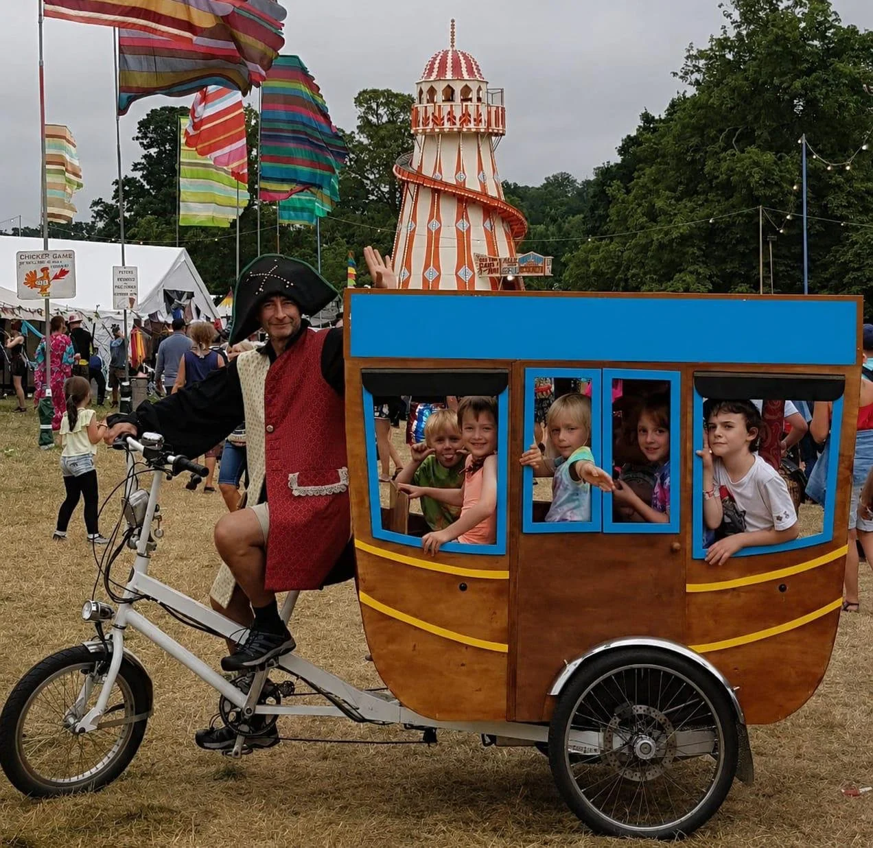 A man dressed as a pirate riding a bicycle with a large wooden carriage attached, designed to look like a pirate ship with children sitting inside, at a fairground with tents, colorful flags, and a circus tent in the background.