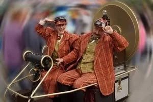 Two people in vintage costumes riding an old-fashioned boat at a parade or event, smiling and holding drinks.