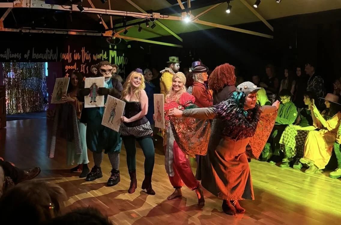 Group of people in colorful costumes performing on stage, some holding paintings, in a theatrical setting with audience watching.
