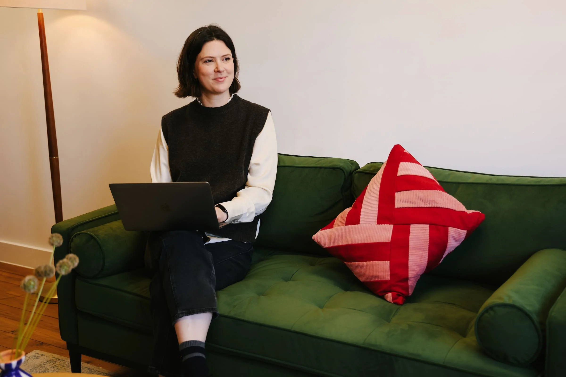 Photo of Claire Buky-Webster sitting on a green sofa with bright red cushion in a warmly-lit South London therapy room