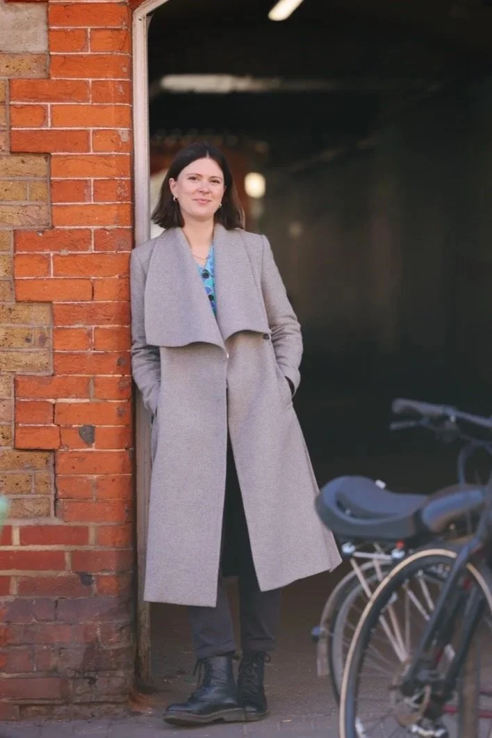 Full length shot of Dr Claire Buky-Webster, South London clinical psychologist and private therapist, outdoors in South London, smartly dressed leaning against brick wall.