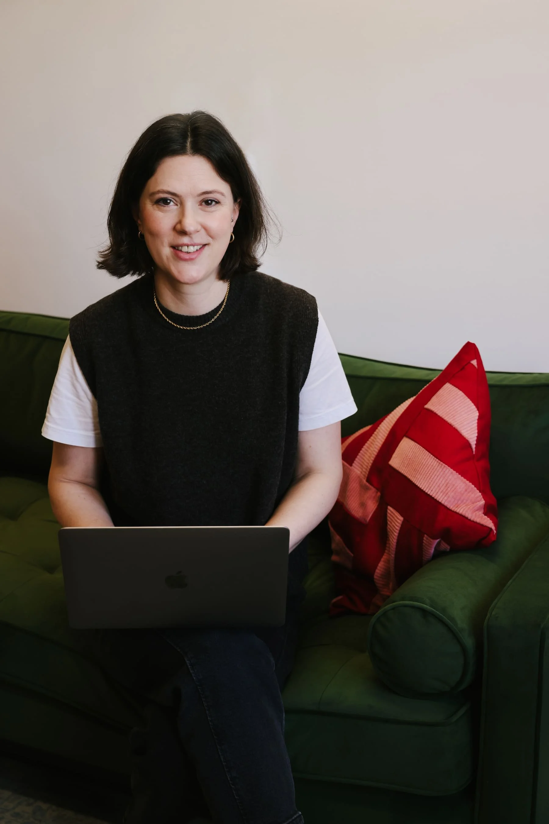 Photo of Claire Buky-Webster sitting on a therapy couch with a laptop and smiling