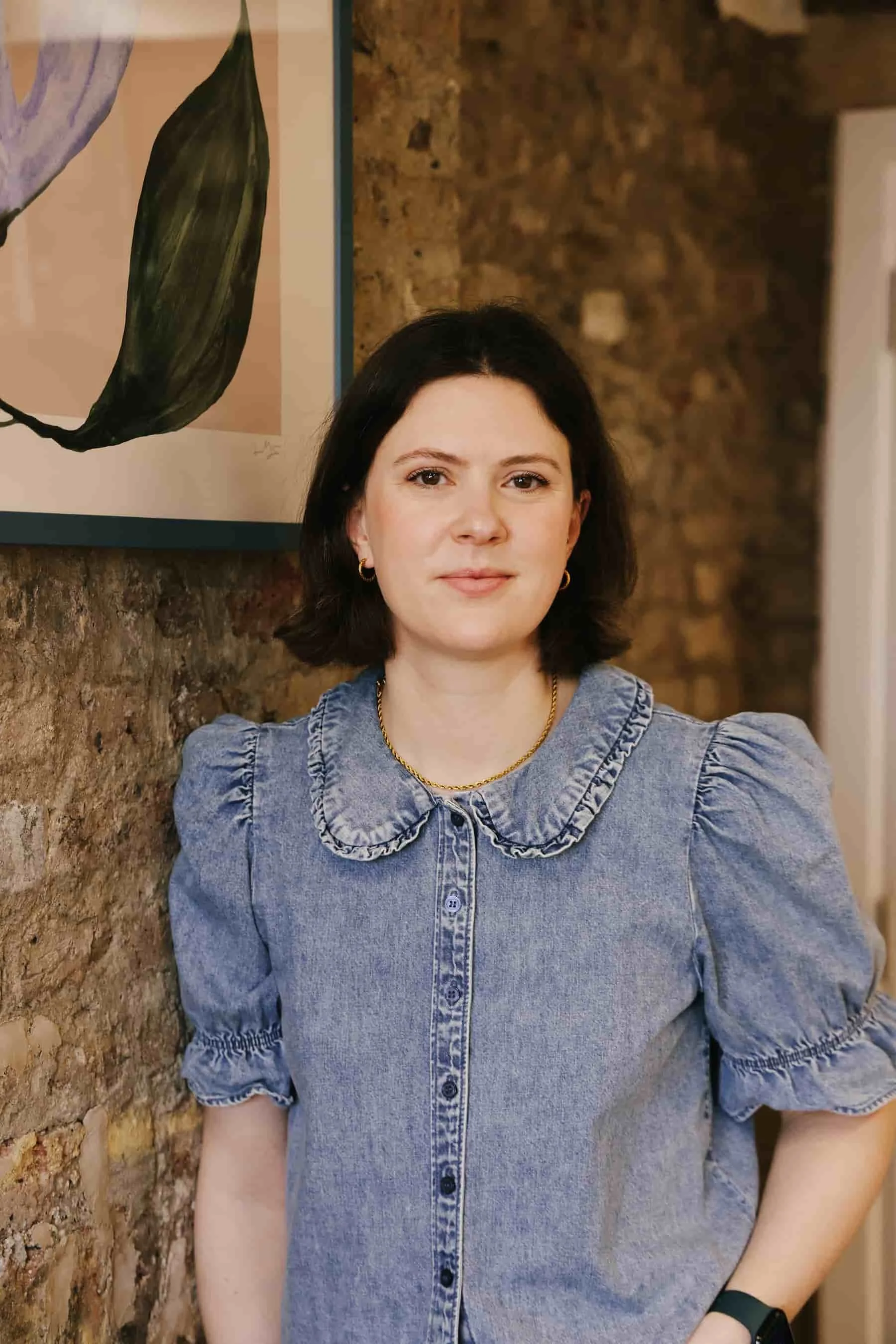 Photo of Claire Buky-Webster, Clinical Psychologist, leaning against an exposed brick wall with a framed botanical print from her therapy room in Brixton