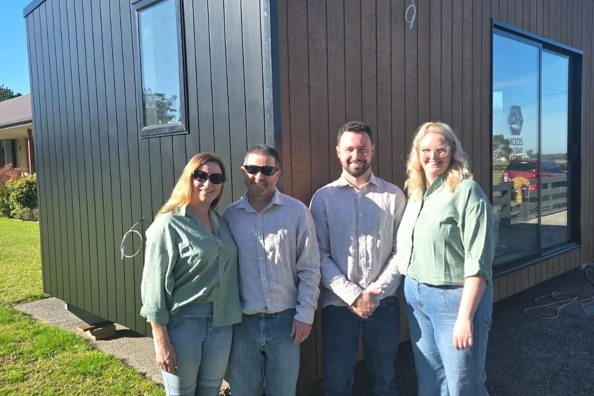Four people standing outside a modern building with large windows, smiling for the photo, on a sunny day.