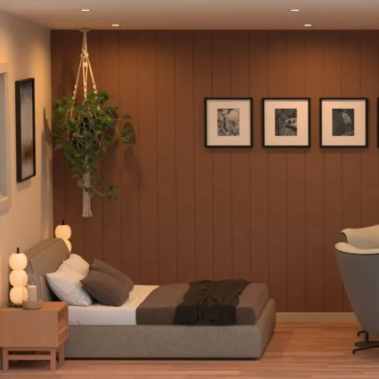 A cozy bedroom with a low bed, beige headboard, and gray bedding. Next to the bed is a wooden nightstand with three stacked lamps. A hanging plant is suspended from the ceiling on the left side. The wall behind the bed is wooden and decorated with three black-and-white framed pictures. To the right, there is a gray armchair.