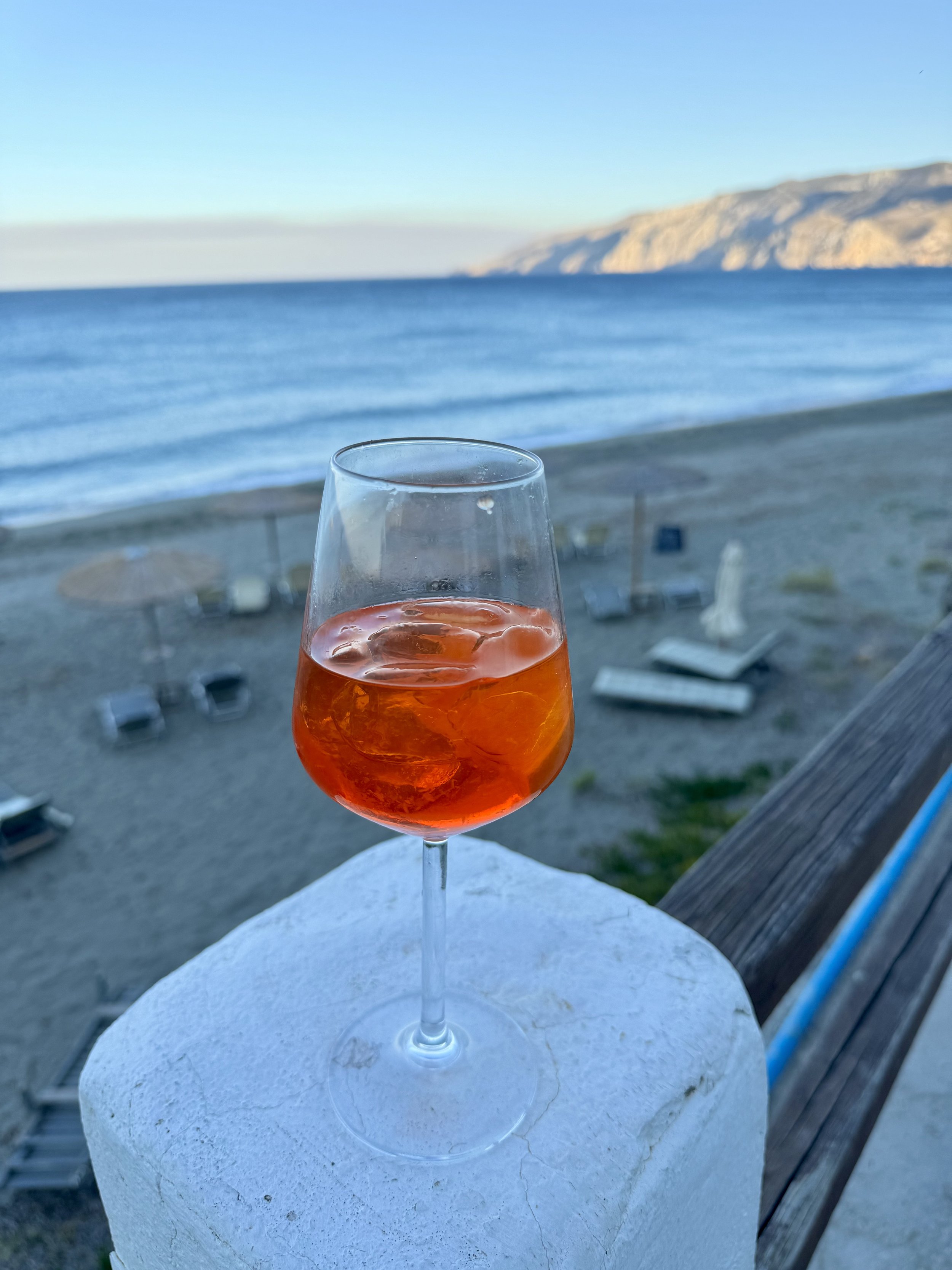 A glass of rosé wine with ice cubes on a white surface overlooking a beach with lounge chairs and umbrellas, calm ocean, and distant mountains under a clear blue sky.