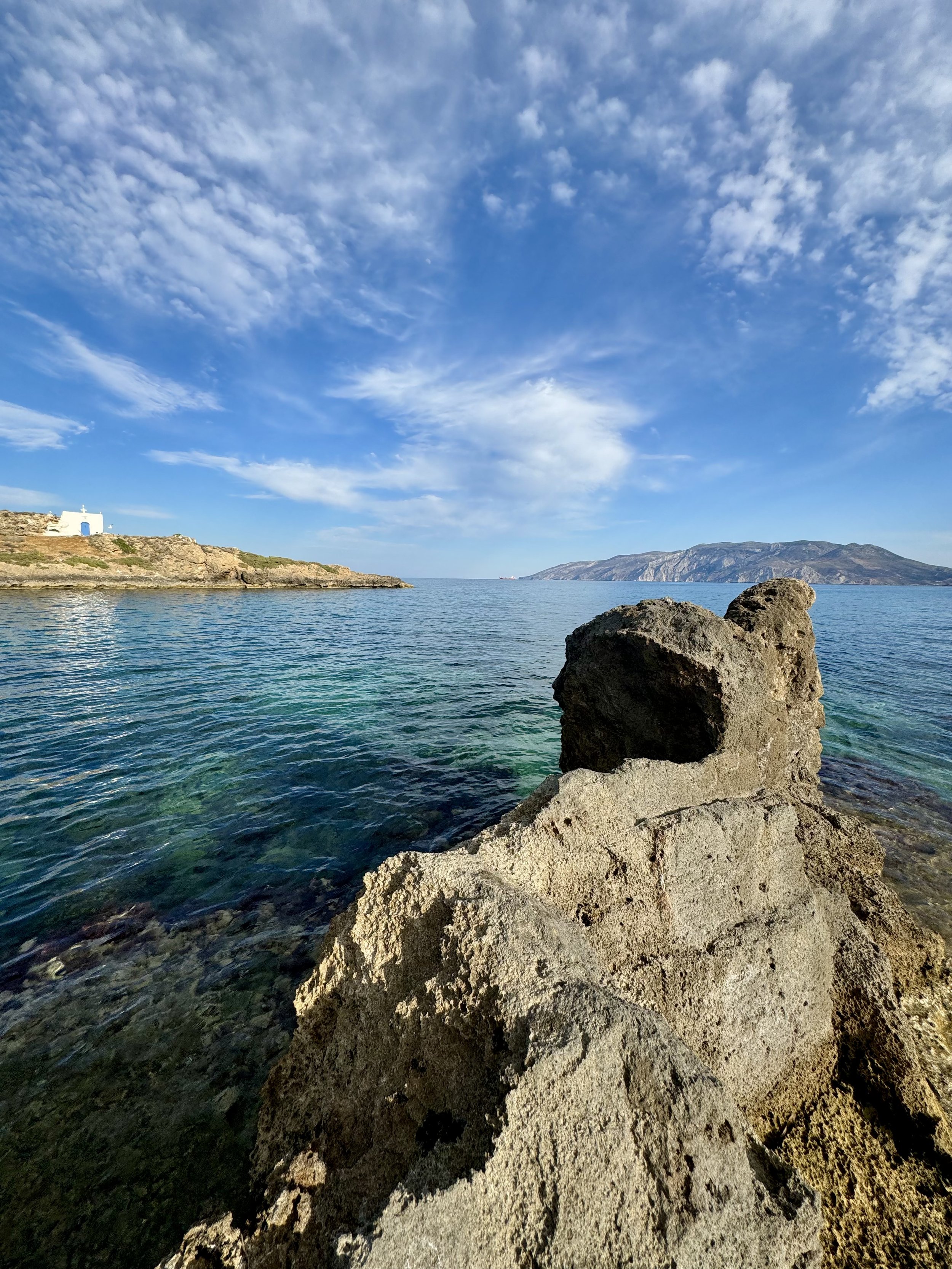 Rocky seaside with clear water, a distant island or mountain, and a small white chapel with a blue door on the coast under a partly cloudy sky.