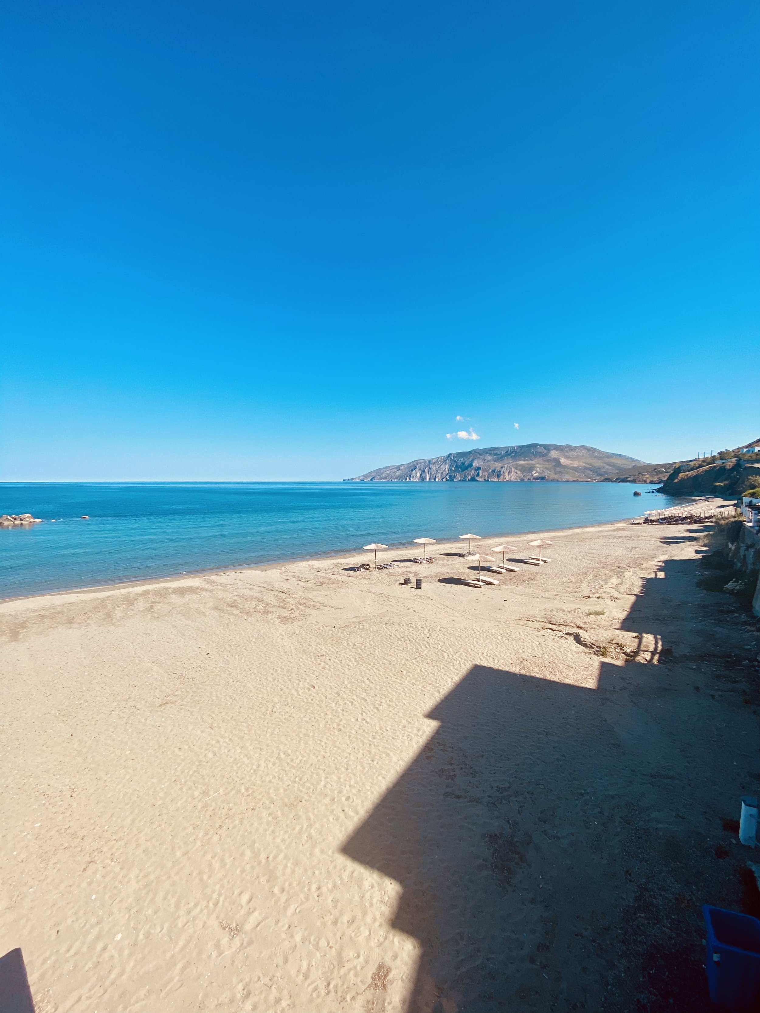 Empty beach with a few umbrellas and lounge chairs, calm blue ocean, distant mountain range, clear blue sky, shadows cast on sandy shore.