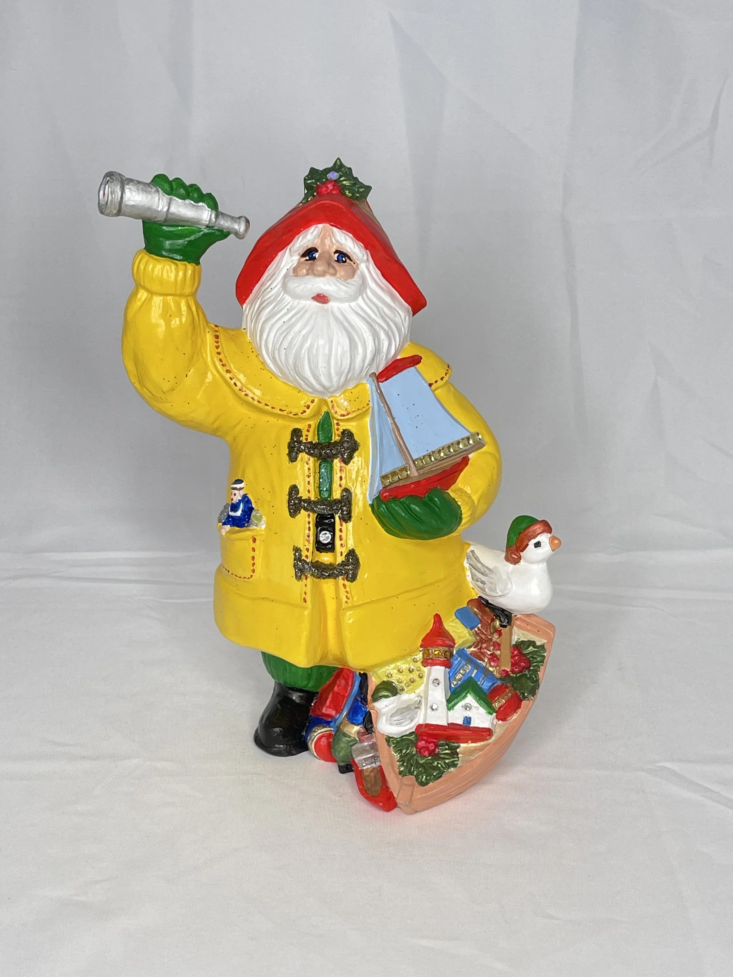 Colorful Christmas figurine of Santa Claus with a white beard, wearing a yellow coat, red hat, and green gloves, holding a silver telescope, with a small sailboat, a white bird, and a decorated boat with a lighthouse and greenery at his feet.
