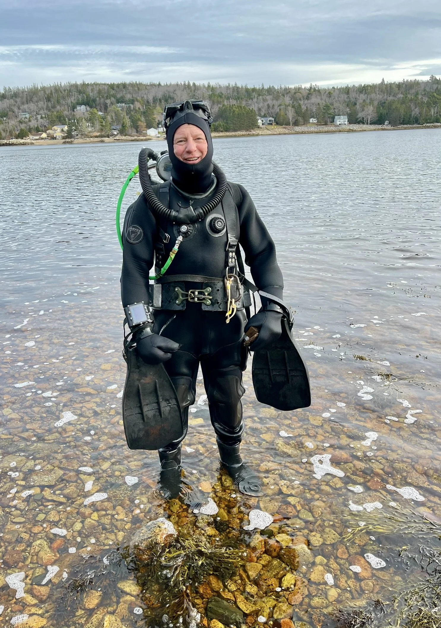 Person in full scuba diving gear standing in shallow water near rocky shoreline, smiling at camera, with houses and trees in the background.