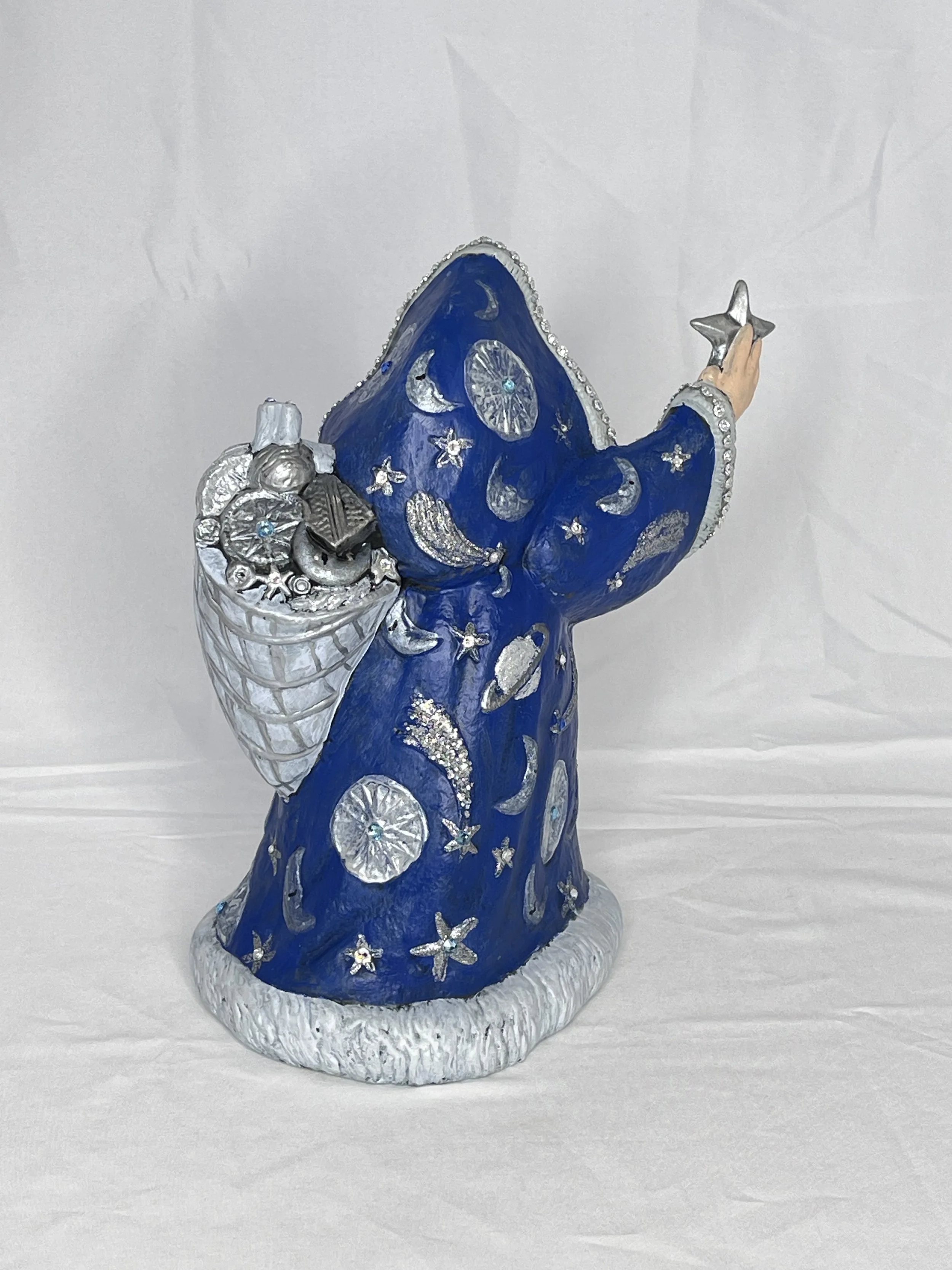 A decorative wizard figure with a blue robe decorated with moons and stars, holding a silver star in one hand and carrying a bag of shiny objects on its back.